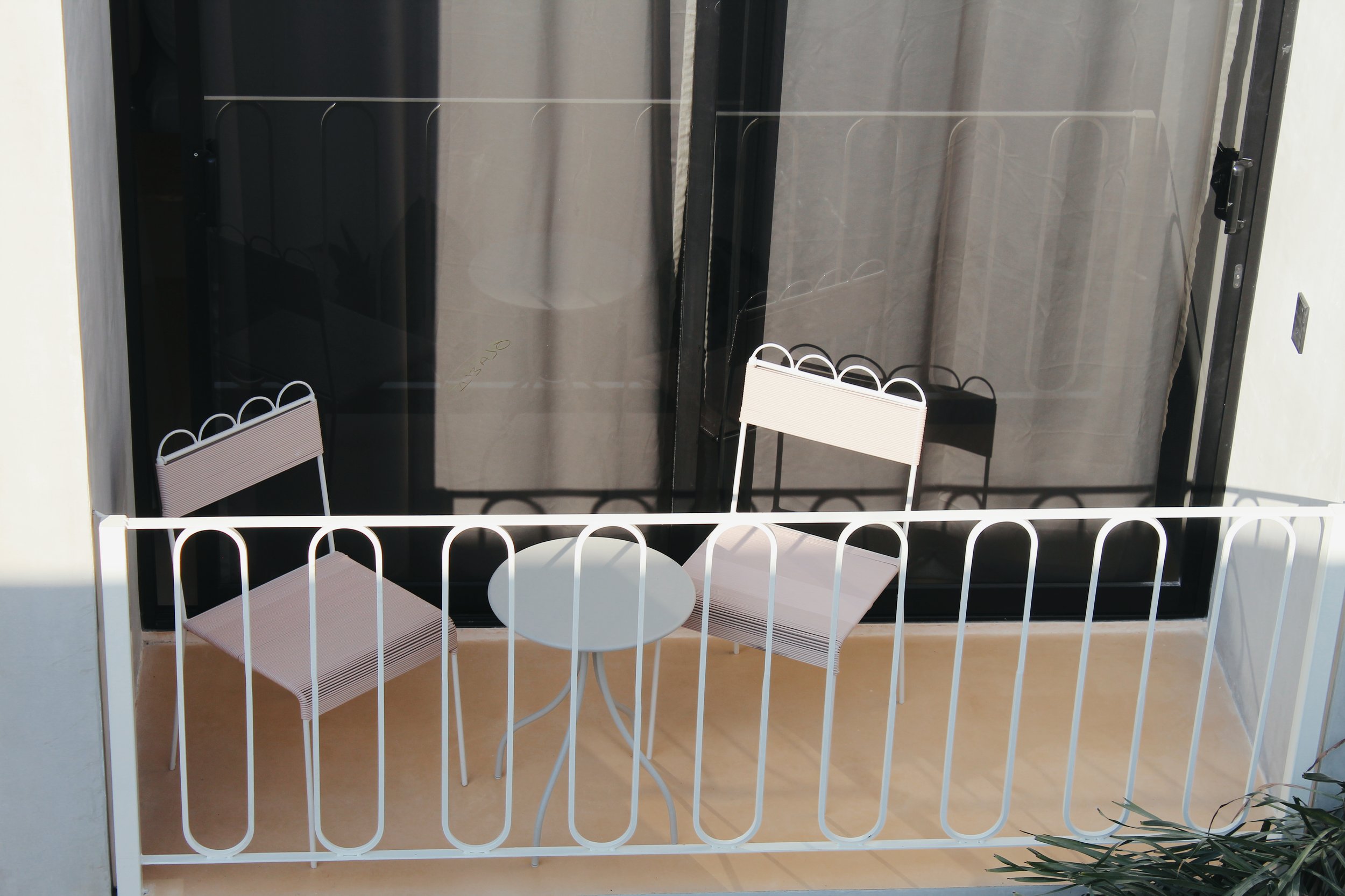 Small balcony with white metal chairs and table, black glass door, and light tan floor.