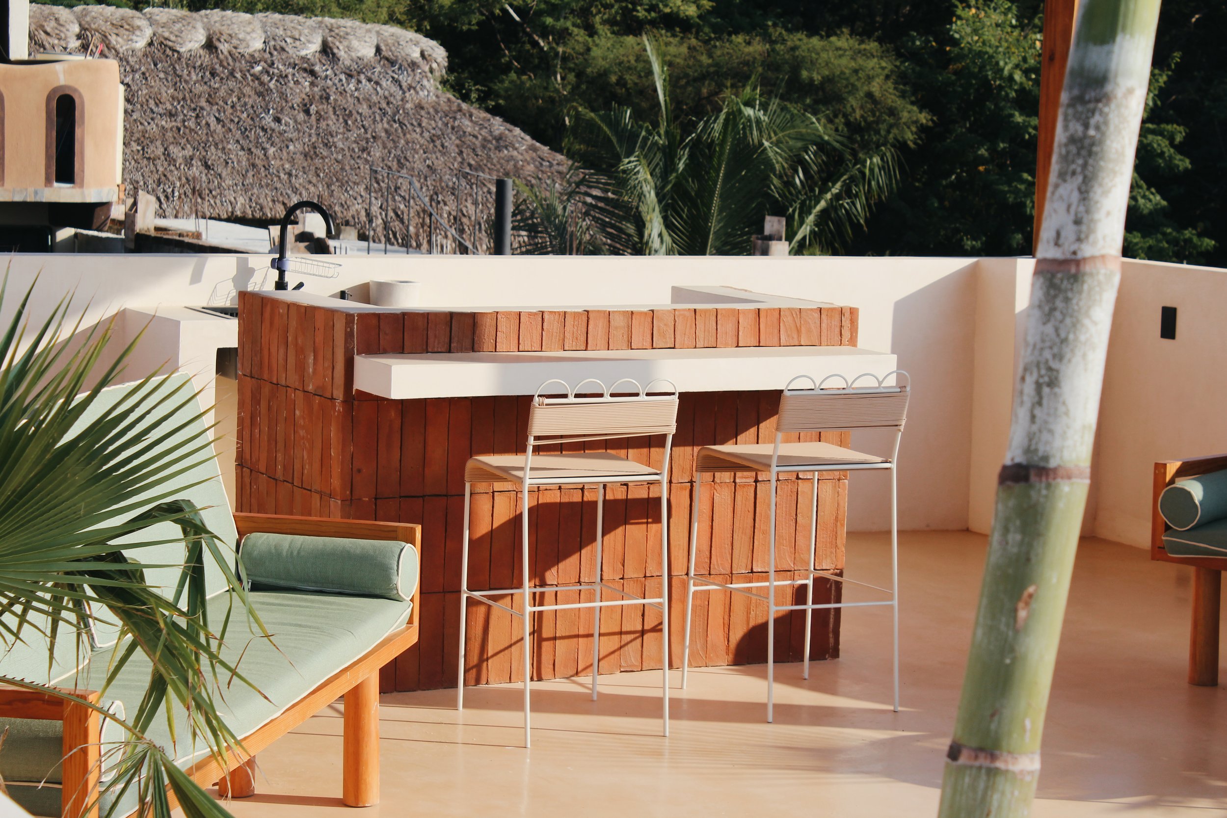 Outdoor patio with brick bar, metal barstools, green cushioned bench, bamboo pole, and tropical greenery.