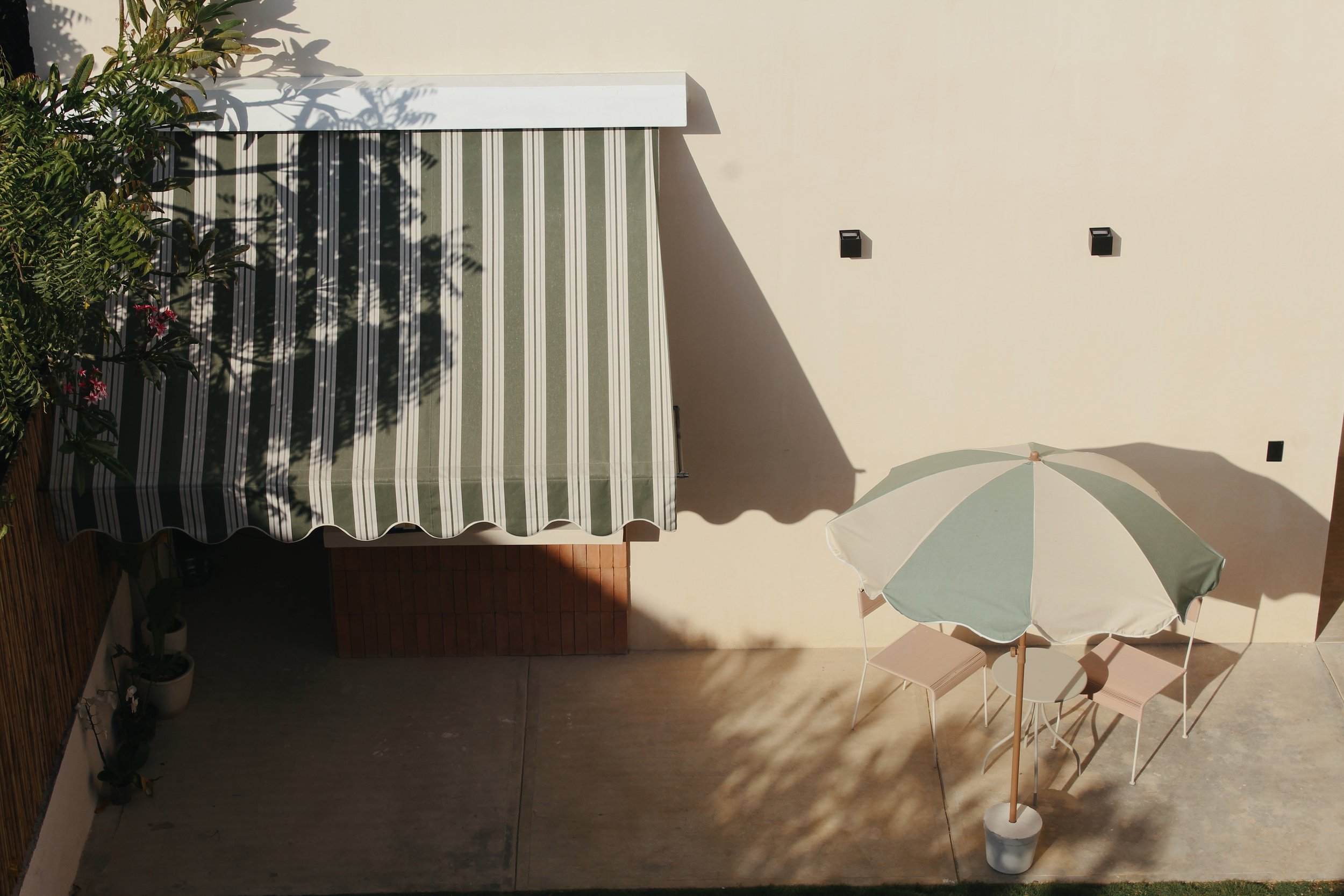 Outdoor patio with green and white striped awning and umbrella, chairs and small table beneath, shadows and a beige wall in background.