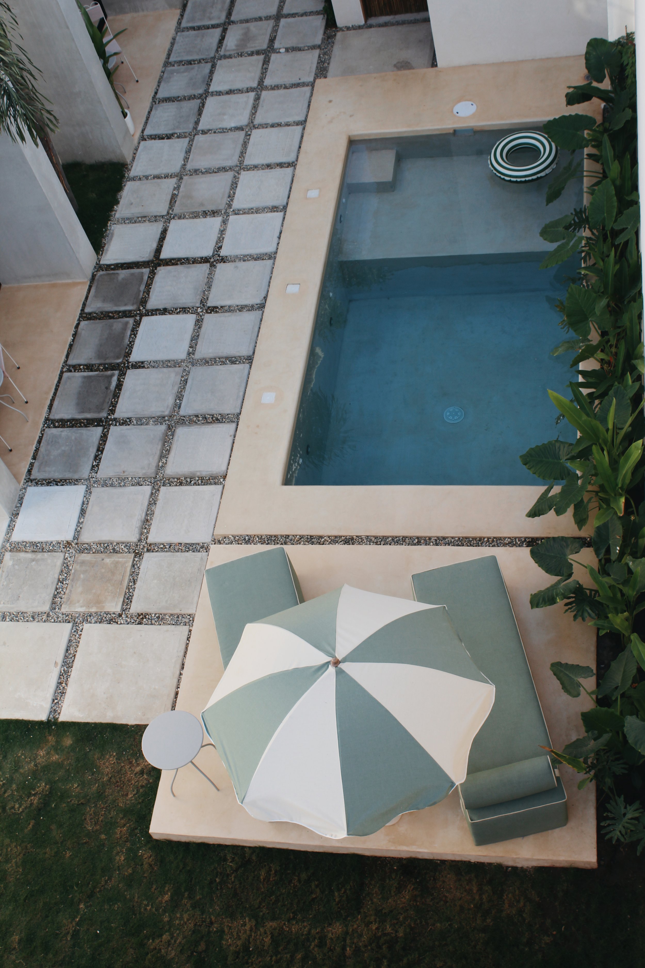 Aerial view of a modern swimming pool next to a walkway with square tiles. There is a green and white striped umbrella over a sunbed and a small round table on a patio area. Surrounding the pool and patio are lush green plants.