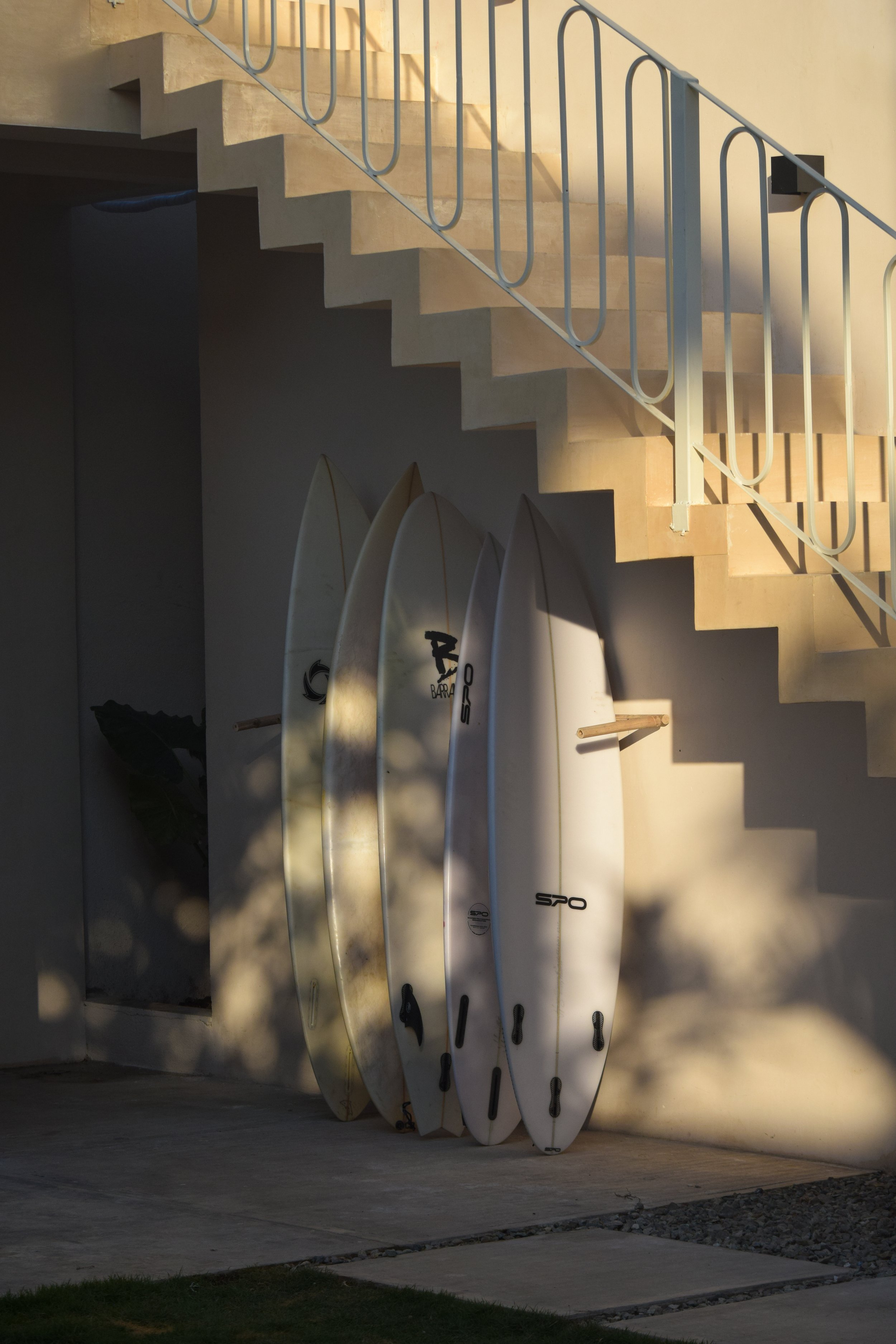 Four surfboards leaning against the wall under a staircase with shadows.