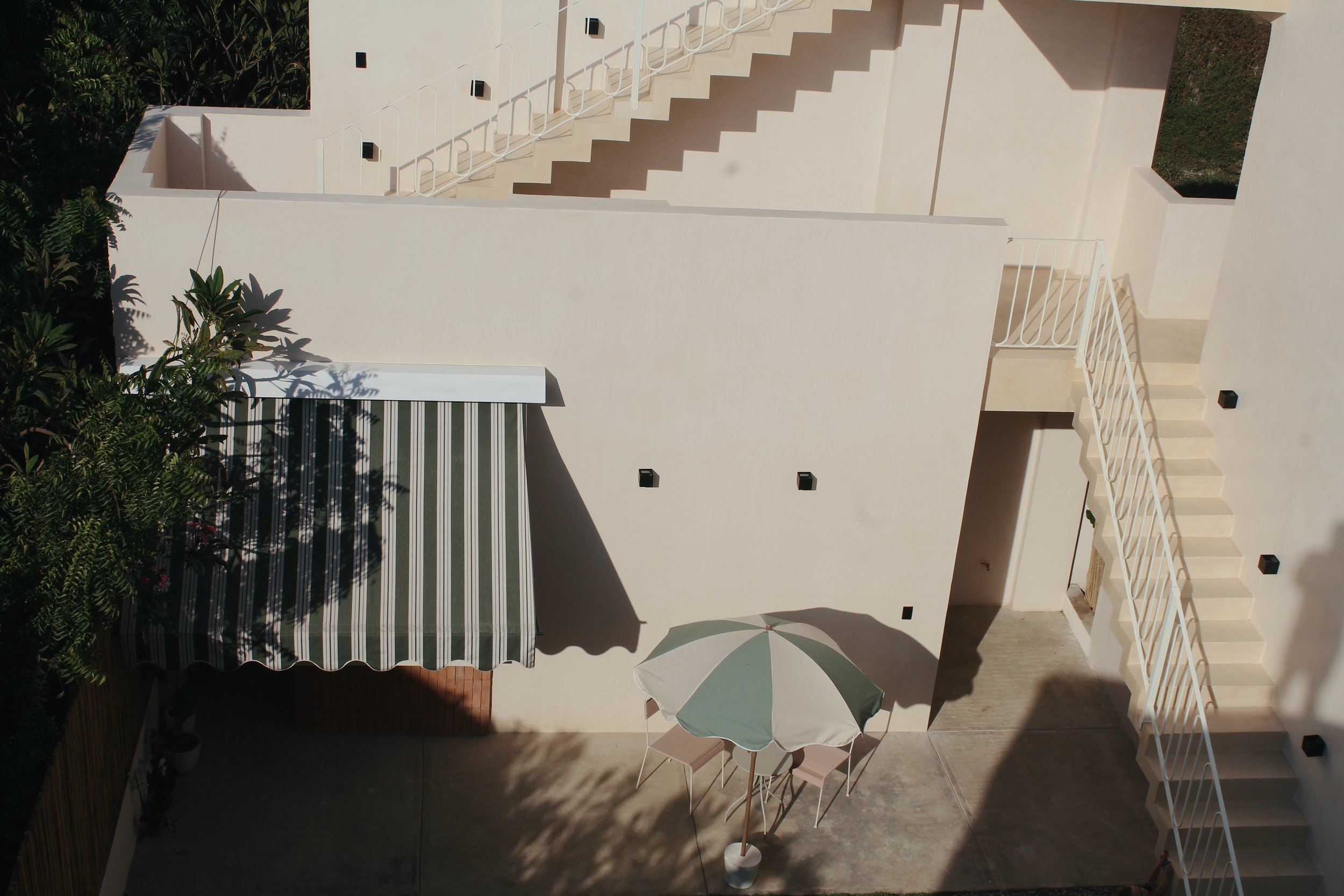 Exterior of a building with cream walls and two sets of stairs. A green and white striped awning and a matching umbrella with chairs are seen on a concrete patio, surrounded by foliage.