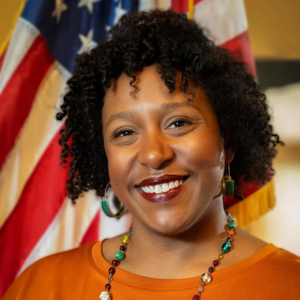 A woman with curly black hair smiling, wearing earrings and a colorful necklace, standing in front of an American flag.