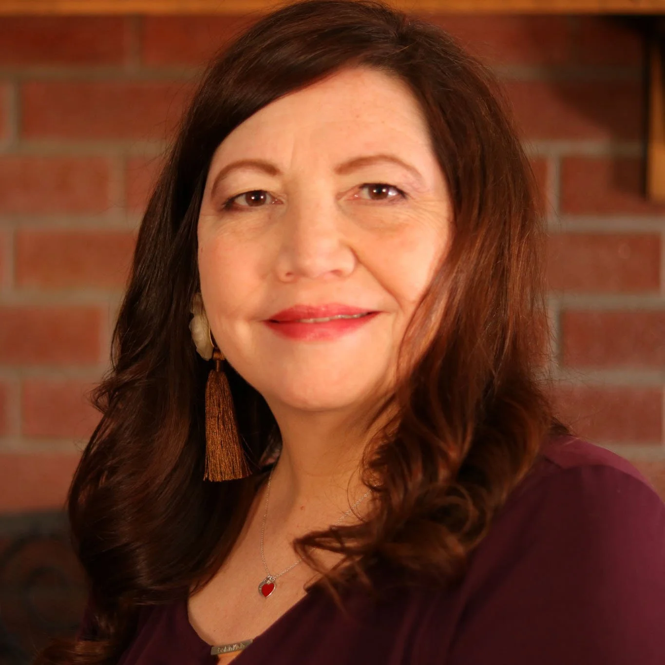 A woman with long, wavy brown hair wearing a maroon top, gold earrings, a silver necklace with a red heart pendant, and a smile, posing in front of a brick wall.