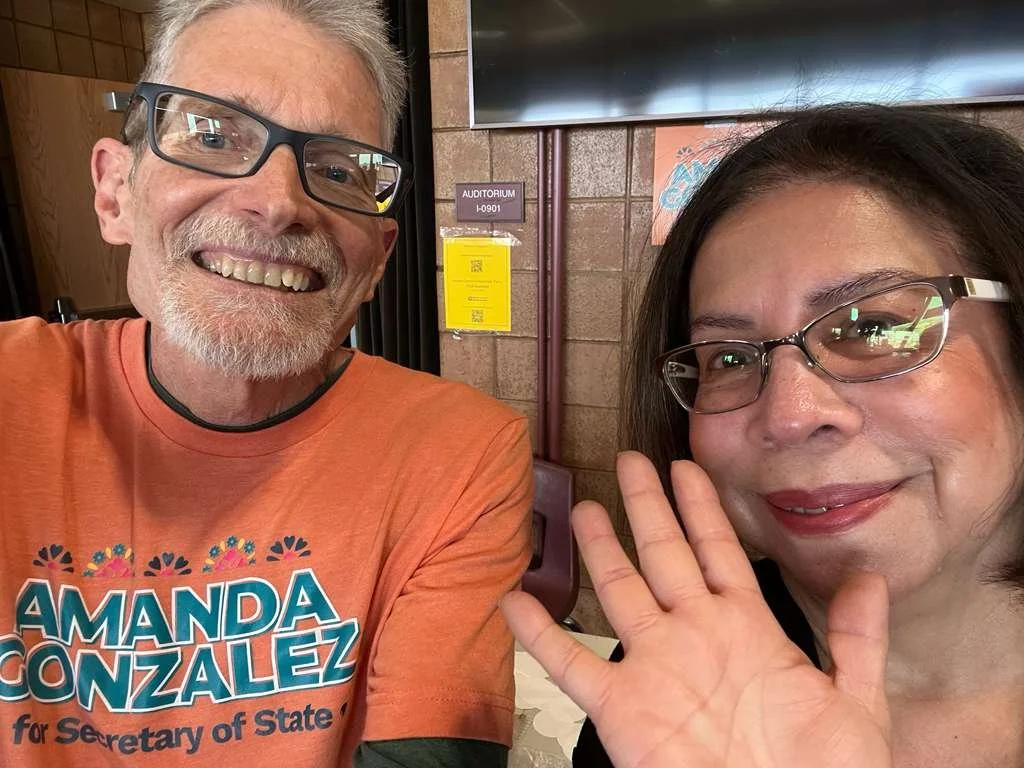 A man and woman taking a selfie indoors. The man wears glasses, a beard, and an orange t-shirt that reads 'Amanda Gonzalez for Secretary of State'. The woman wears glasses, has shoulder-length dark hair, and is waving at the camera.