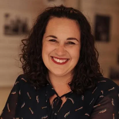 A woman with curly dark hair smiling and wearing a dark blouse with a pattern, indoors in front of a blurred background.