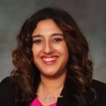Portrait of a woman with curly hair, smiling, wearing a black blazer and pink blouse.