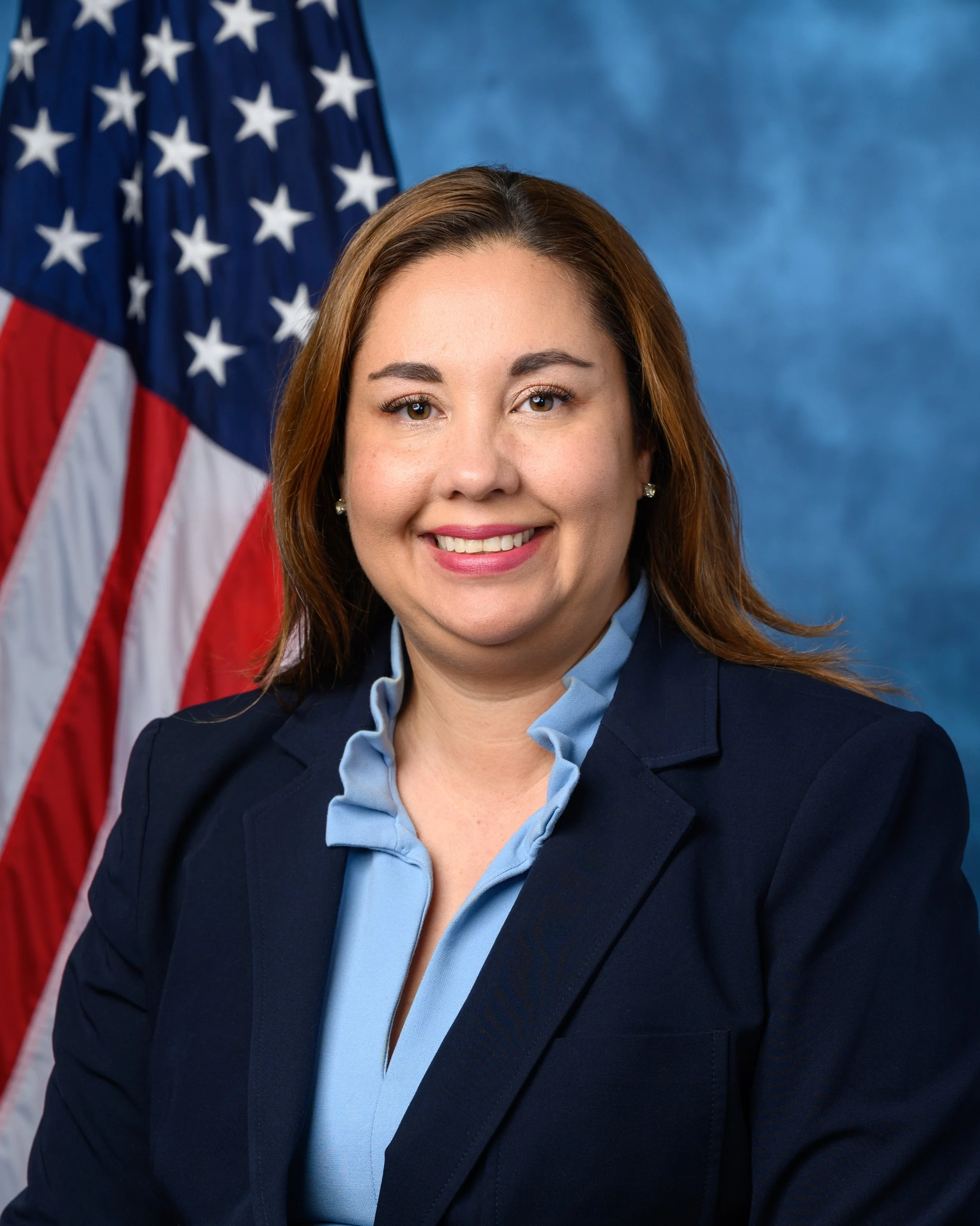 Yadira Caraveo photo, woman with shoulder length brown hair smiles in front of an American flag.