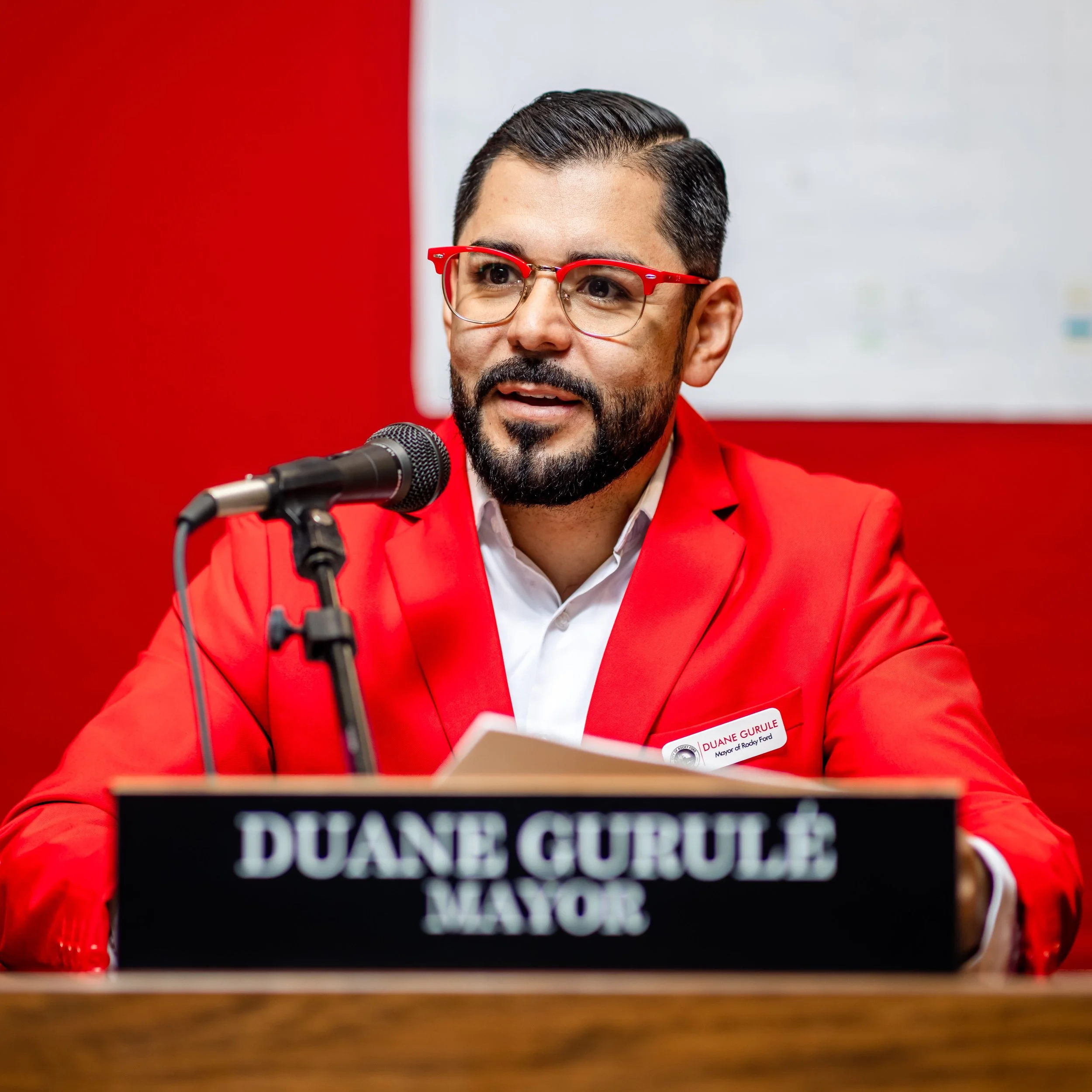 A man with dark hair, beard, glasses with red frames, wearing a red jacket and white shirt, speaking into a microphone at a press conference table with a sign that reads 'DUANE GURULE MAYOR'.