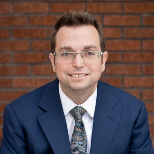 Professional man in a suit with glasses smiling against a brick wall background.