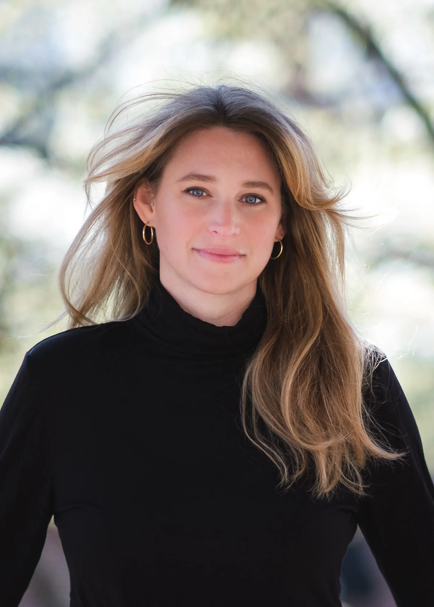 A woman with long wavy hair wearing a black turtleneck sweater, gold hoop earrings, and small earrings, standing outdoors with a blurred background of trees and daylight.