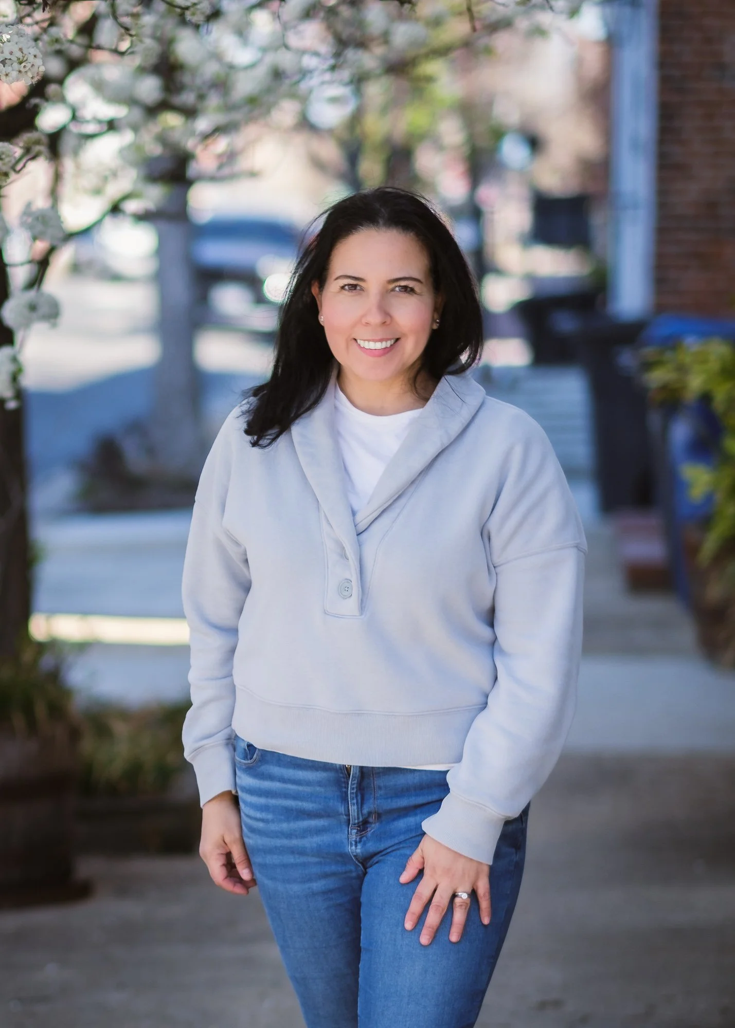 A smiling woman with dark hair wearing a light gray hoodie and blue jeans standing outdoors on a sidewalk with trees and buildings in the background.