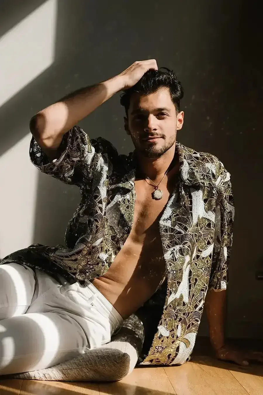 Ein junger Mann sitzt auf dem Boden, trägt ein offen getragenes, florales Hemd, weiße Hosen, hat dunkles Haar und trägt eine Halskette. Er ist bei Sonnenlicht, das Schatten auf die Wand hinter ihm wirft, und lächelt leicht.