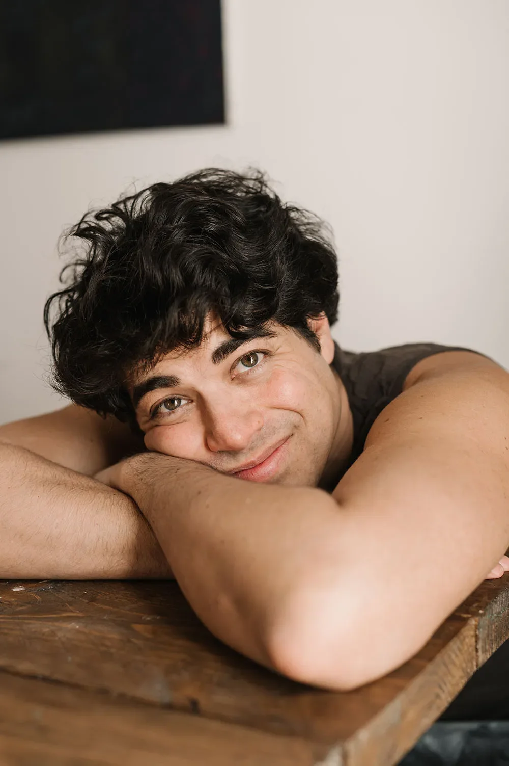 Junger Mann liegt auf einem Holztisch, kuschelt mit seinem Kopf auf seinen Armen und lächelt freundlich in die Kamera.