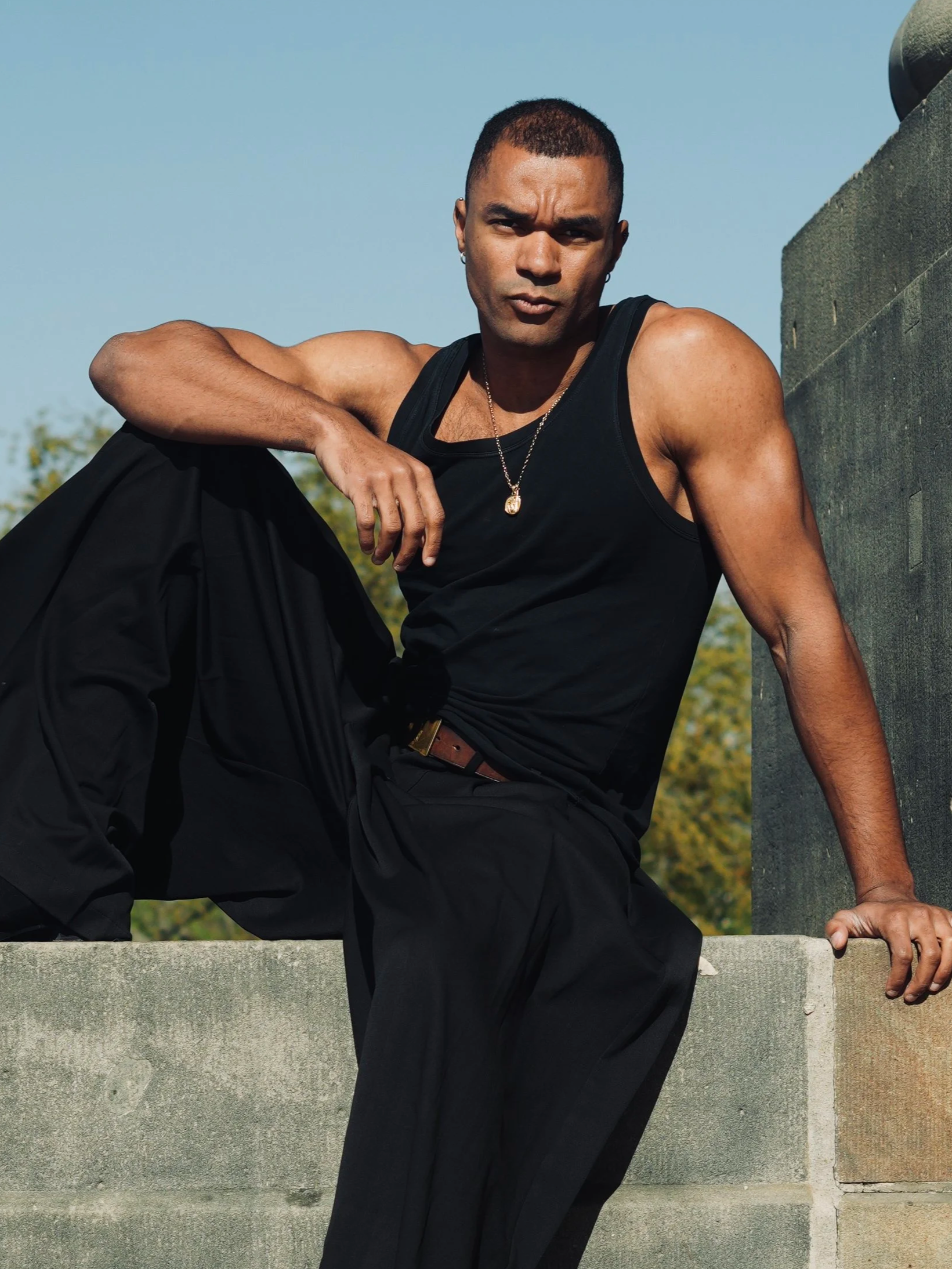 A man with short hair and earrings dressed in a black tank top and pants, sitting on a concrete structure outdoors during daytime, leaning against a wall with a serious expression.