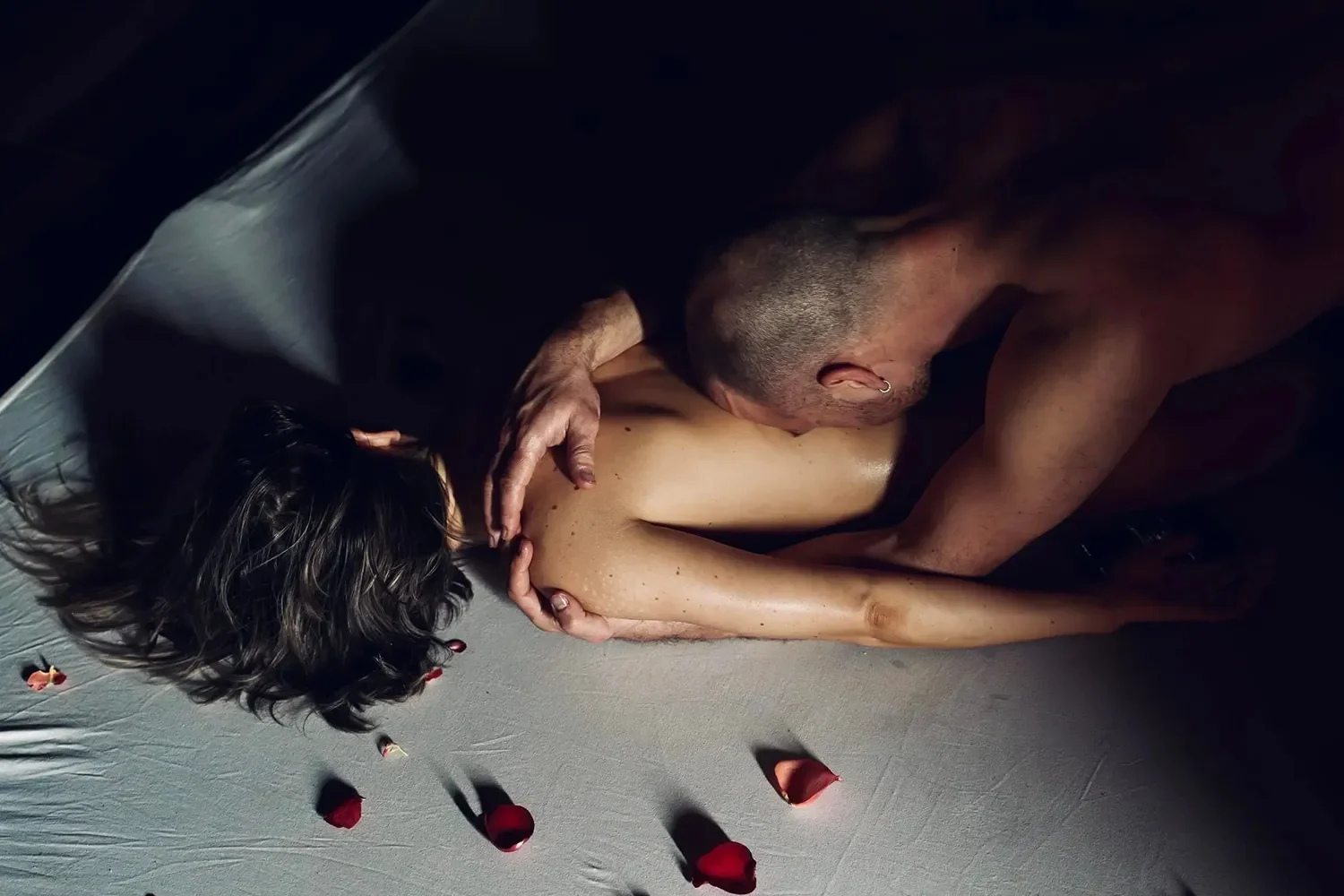 A man and woman engaged in an intimate embrace on the floor, with rose petals scattered around them.
