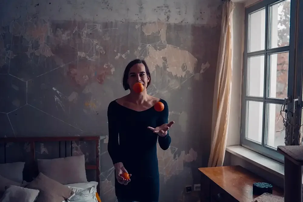 A woman in a dark dress is inside a room with a bed and a wooden table, balancing three orange balls in the air, two on her shoulders and one near her mouth, with a window to her right.