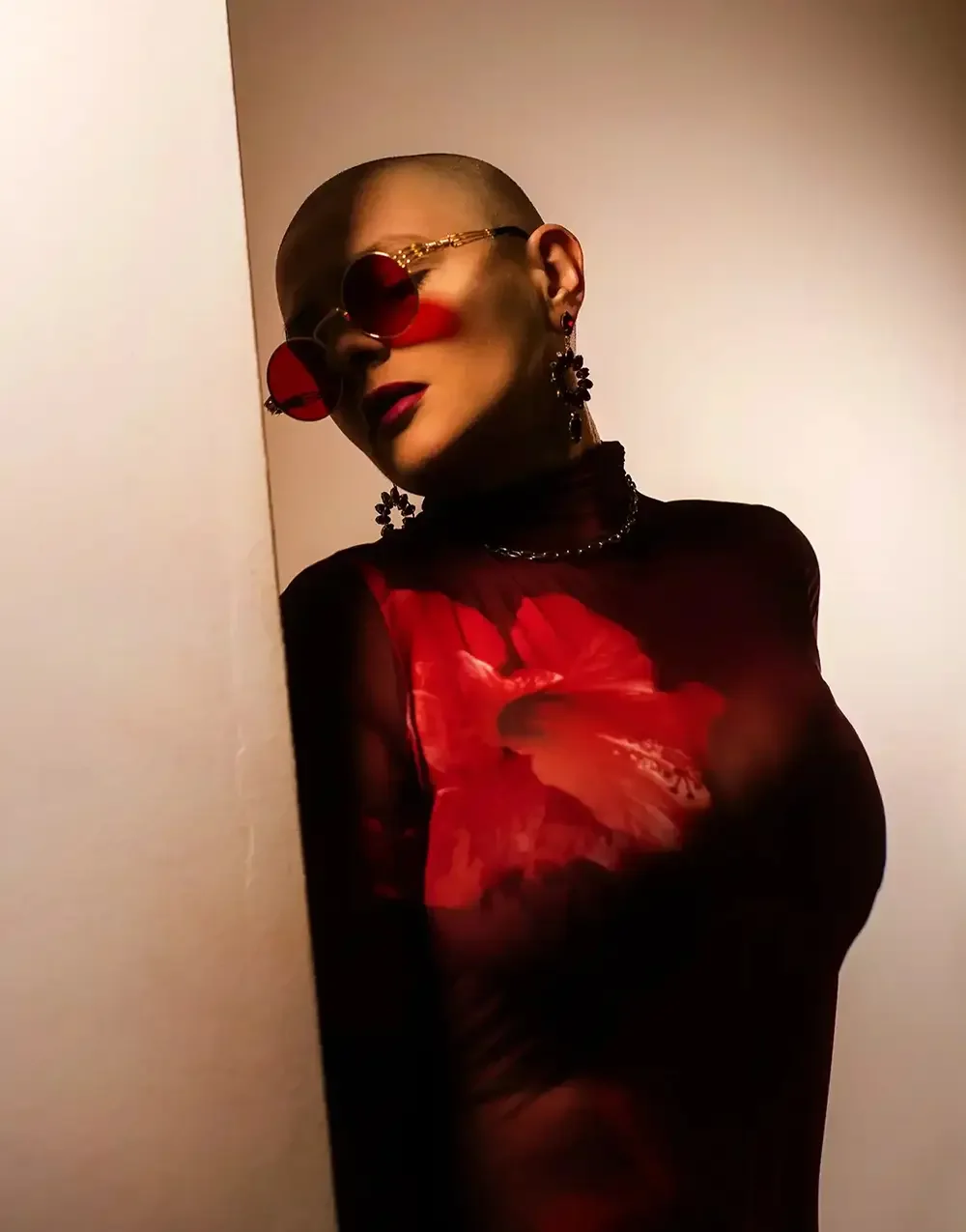 A person with a shaved head wearing red-tinted sunglasses, black floral earrings, a black choker, and a black sheer top with a red floral pattern, posing against a plain background.
