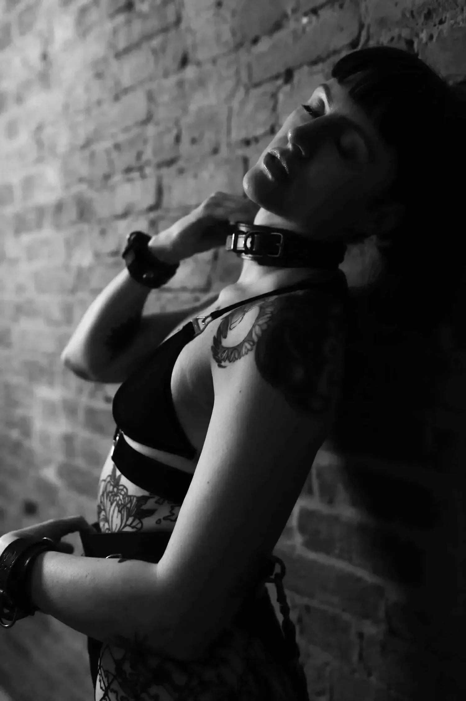 A black and white photo of a tattooed woman with short hair, wearing a collar and a sexy top, leaning against a brick wall.