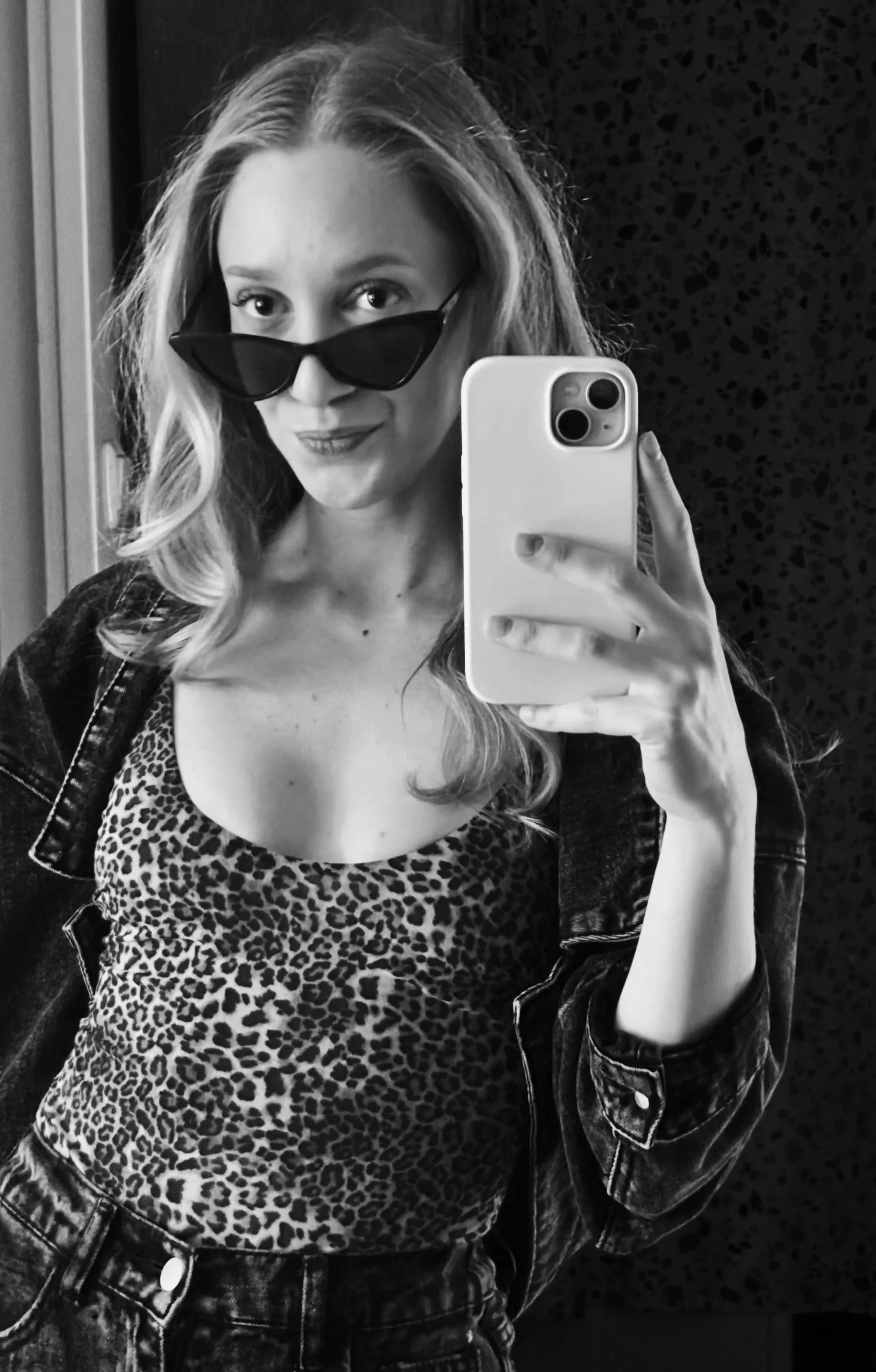 Black and white selfie of a woman with wavy hair, wearing sunglasses, a leopard print top, and a denim jacket, holding a smartphone in front of a mirror.