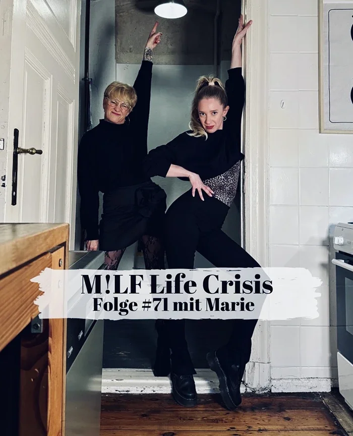 Two women posing inside a kitchen, one older with glasses and a black outfit, and one younger with a ponytail and black outfit, standing on tiptoe and striking a pose.