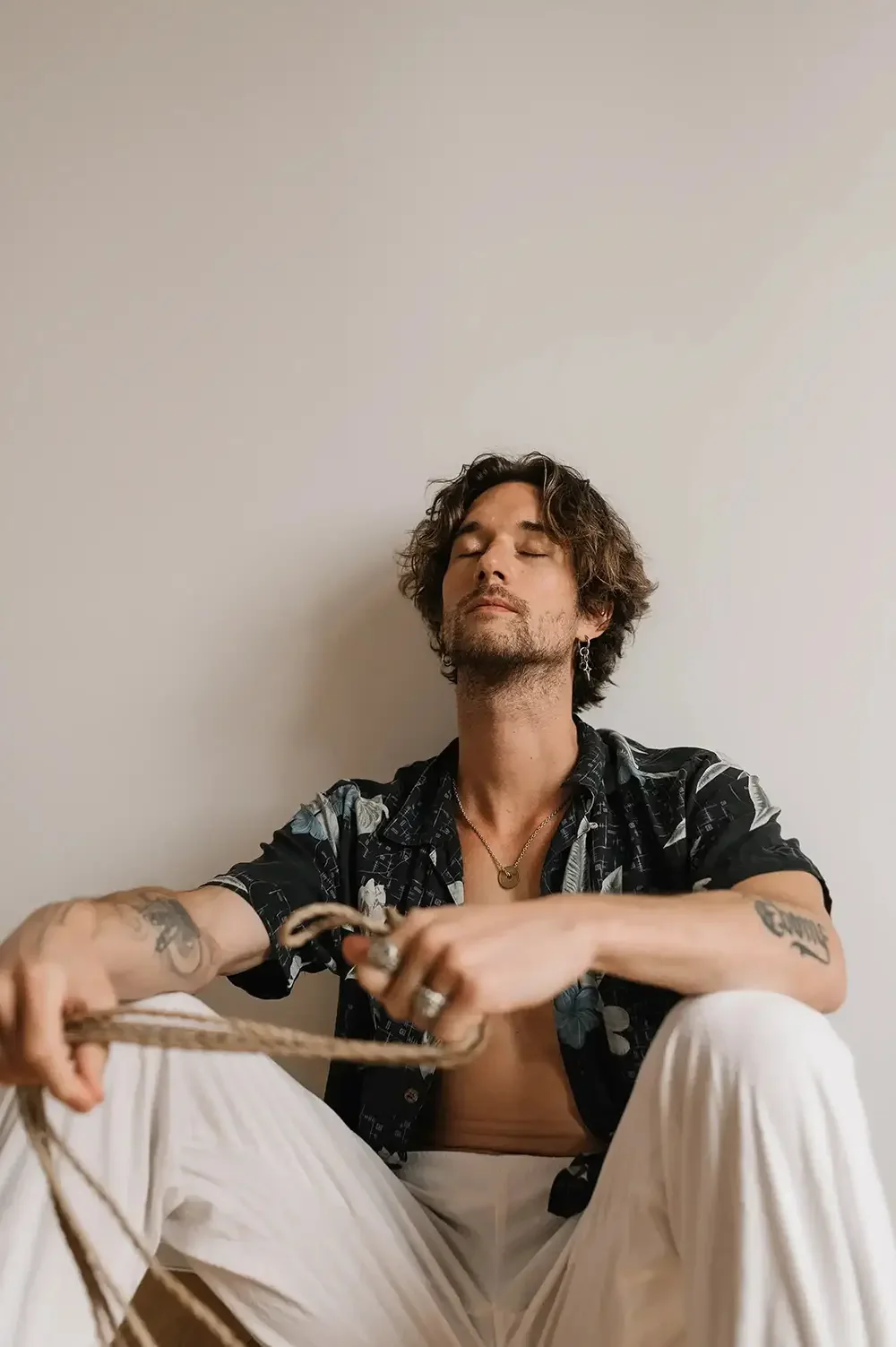 A man with tattoos and jewelry sitting against a plain white wall with eyes closed, appearing relaxed.