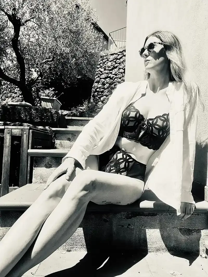 A woman with long hair wearing sunglasses, a white jacket, and a patterned black bikini, sitting outdoors with her legs extended, shadowed by sunlight.