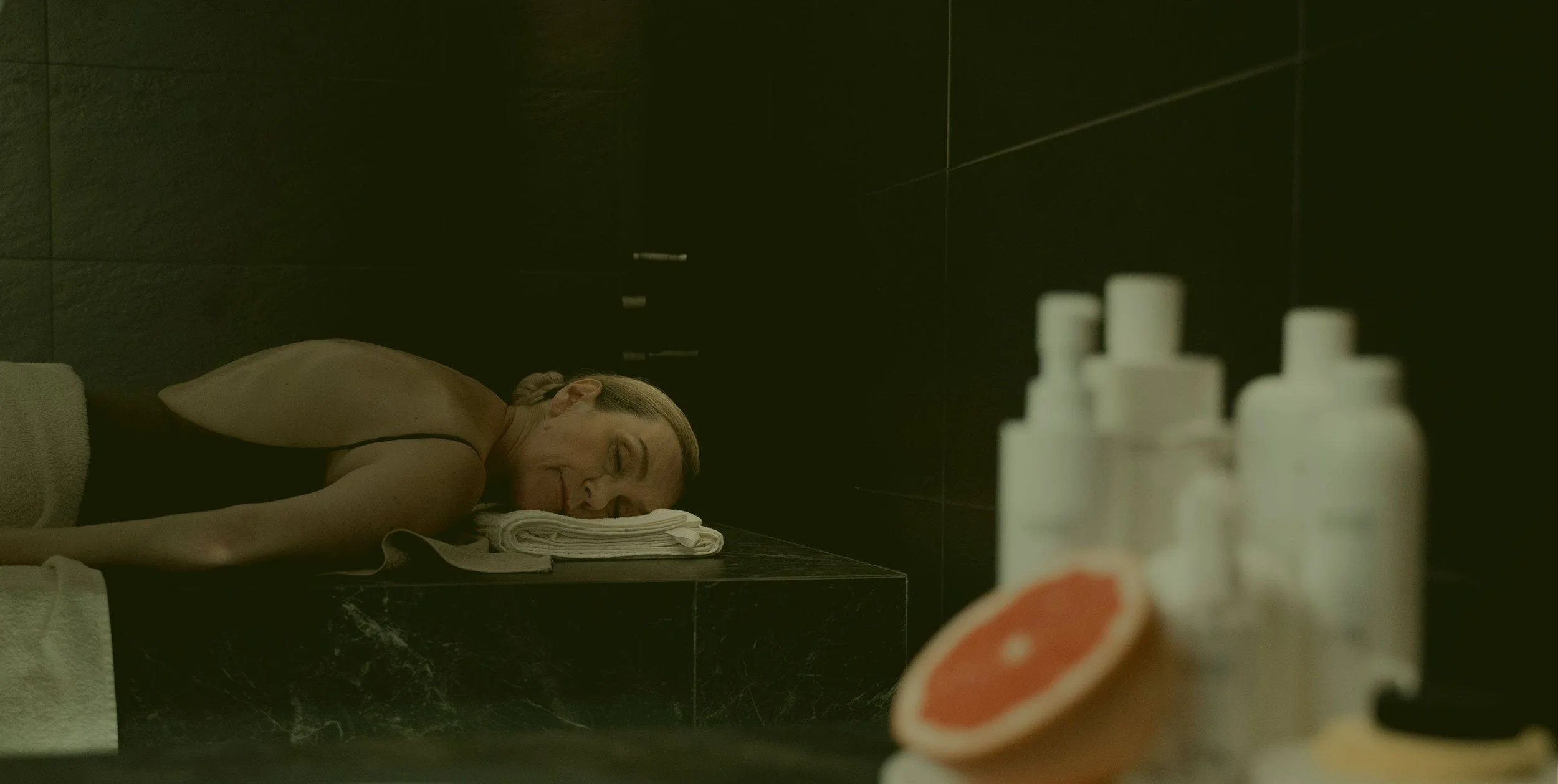 Older lady relaxing on a massage table in a sauna room, with a blurred background and orange-colored massage lotions in the foreground, creating a serene and tranquil spa atmosphere.