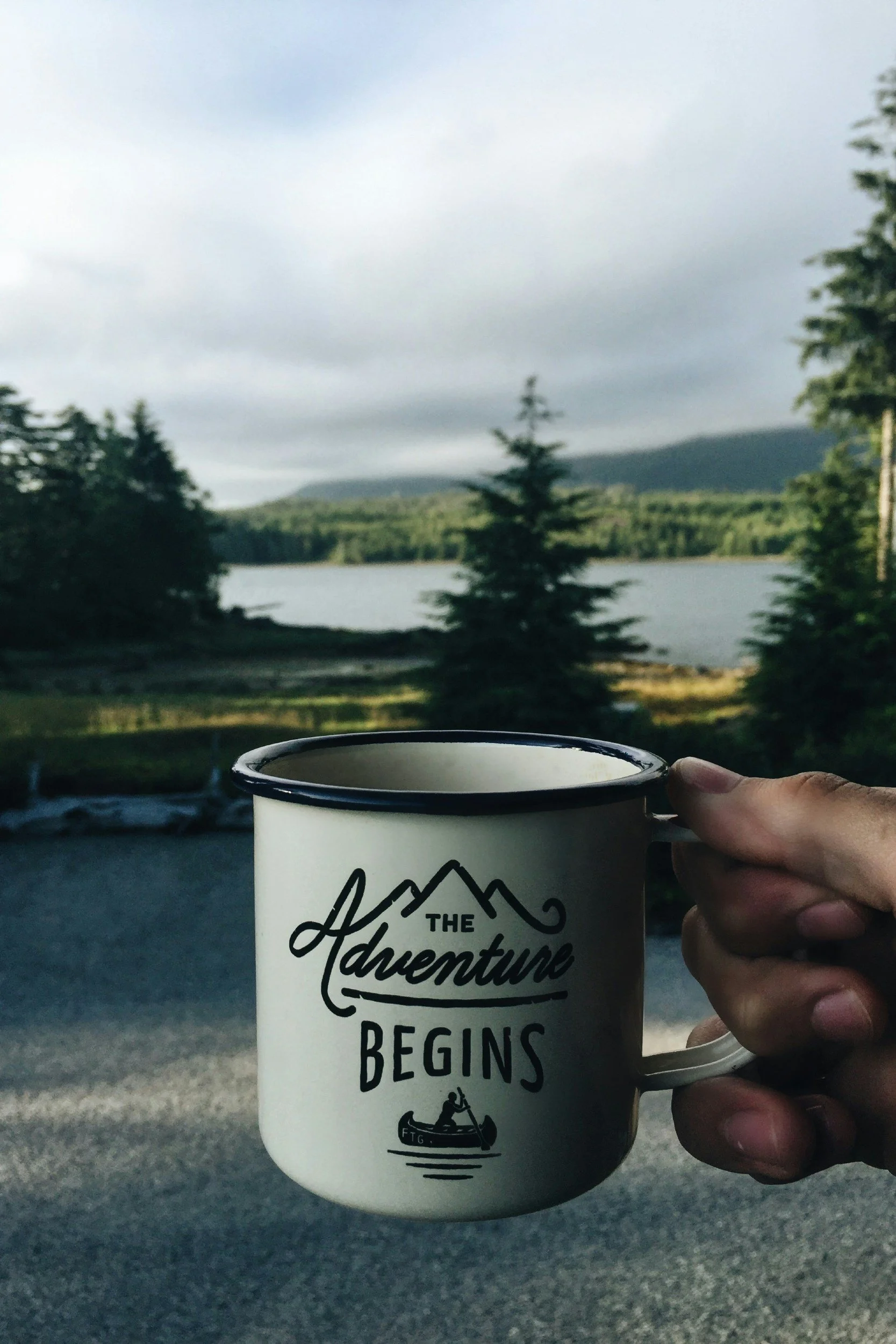 Hand hält Tasse mit der Aufschrift 'The Adventure Begins' vor einer naturschönen Landschaft mit Bäumen und einem See im Hintergrund.