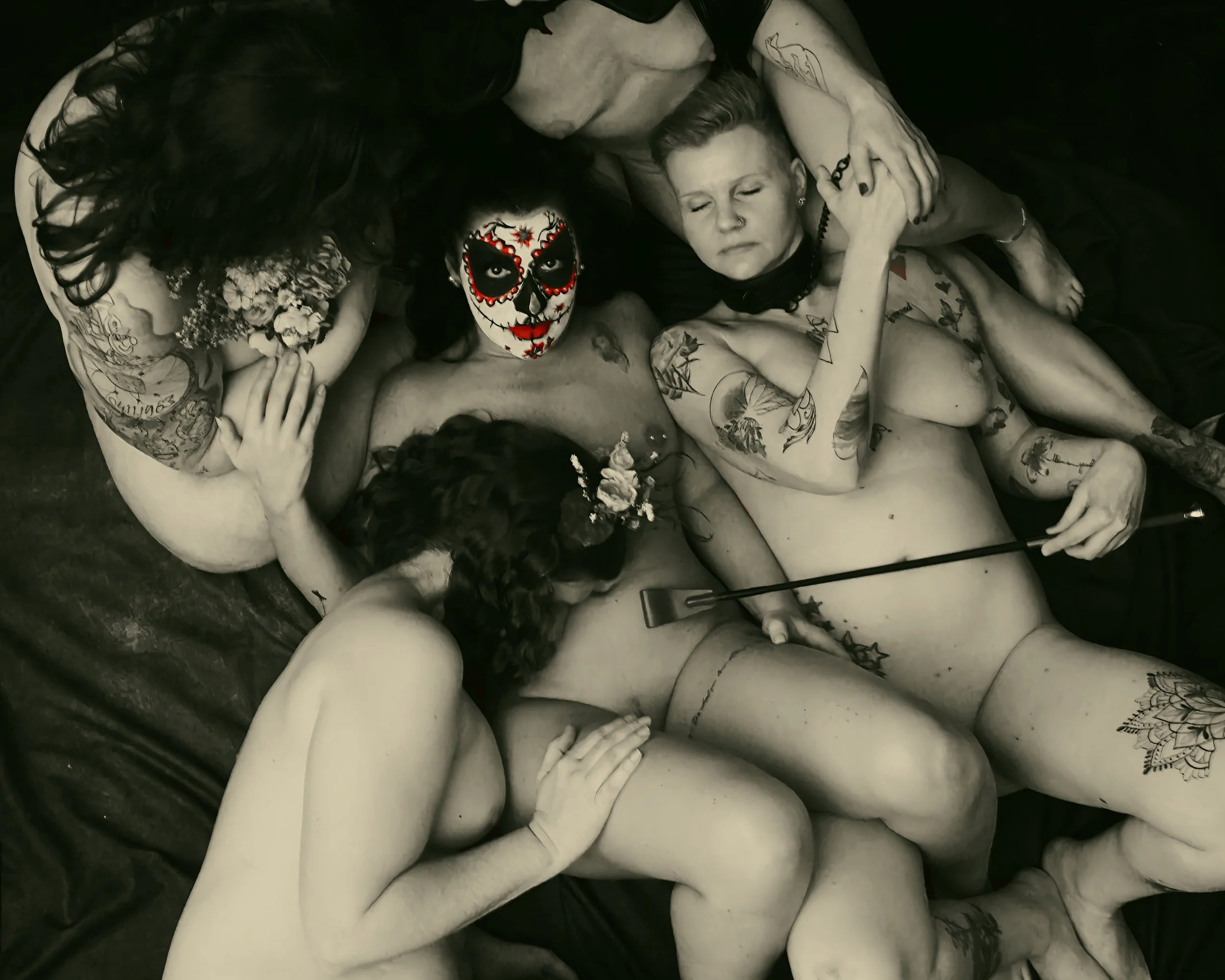 Eine Gruppe tätowierter Frauen, die eng beieinander liegen. Eine Frau trägt eine bunte Schädel-Maske. Schwarz-weiß Fotografie.