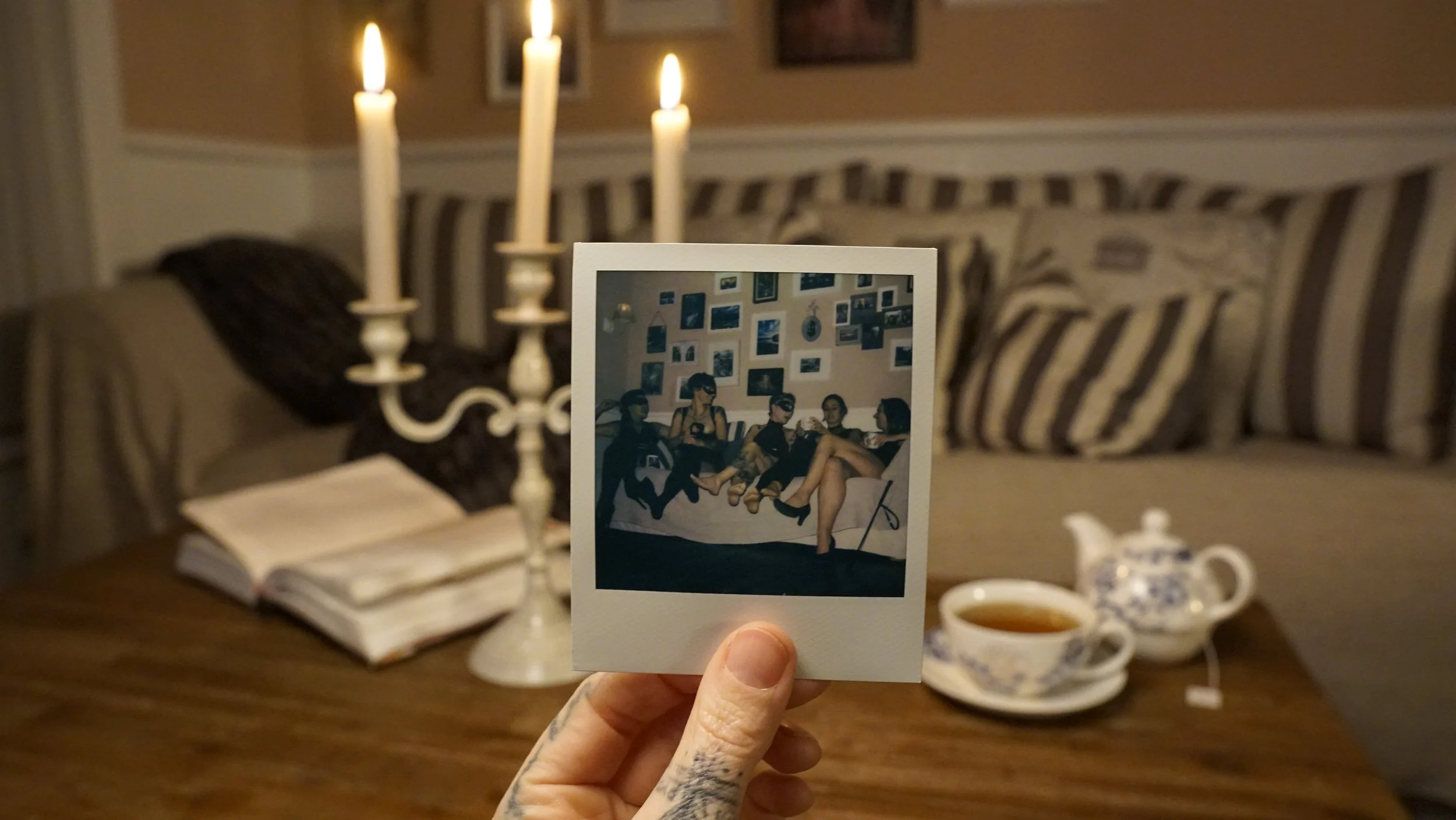 Ein hell erleuchteter Raum mit gemütlicher Einrichtung. Eine Hand hält ein Polaroid, das eine Gruppe von Menschen auf einem Sofa zeigt. Im Hintergrund stehen brennende Kerzen auf einem Kandelaber, Bücher liegen auf einem Tisch, und ein Tee-Set ist ebenfalls sichtbar.