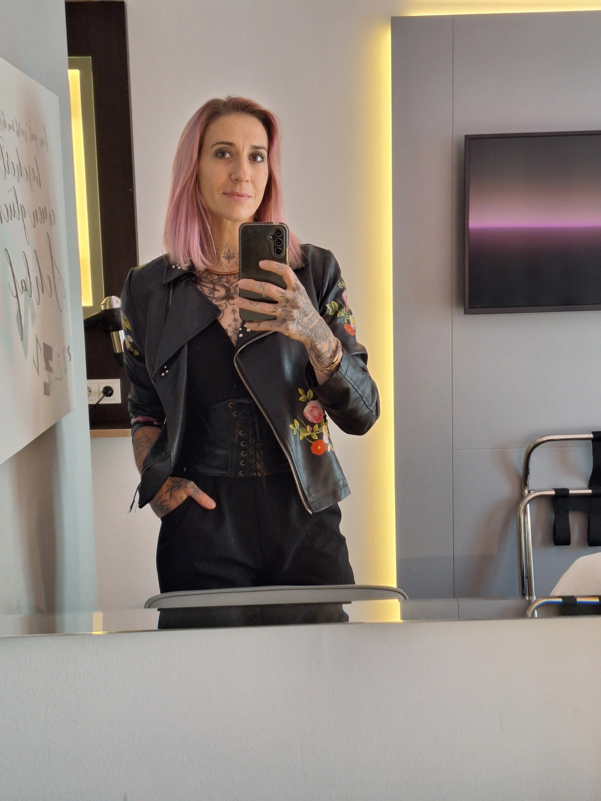 Frau mit pinkfarbenem Haar macht ein Selfie in einem Spiegel. Sie trägt eine dunkle Lederjacke mit Blumenmustern und zeigt Tätowierungen auf ihren Händen und am Hals. Im Hintergrund ist ein Teil eines Raums mit einem Fernseher sichtbar.