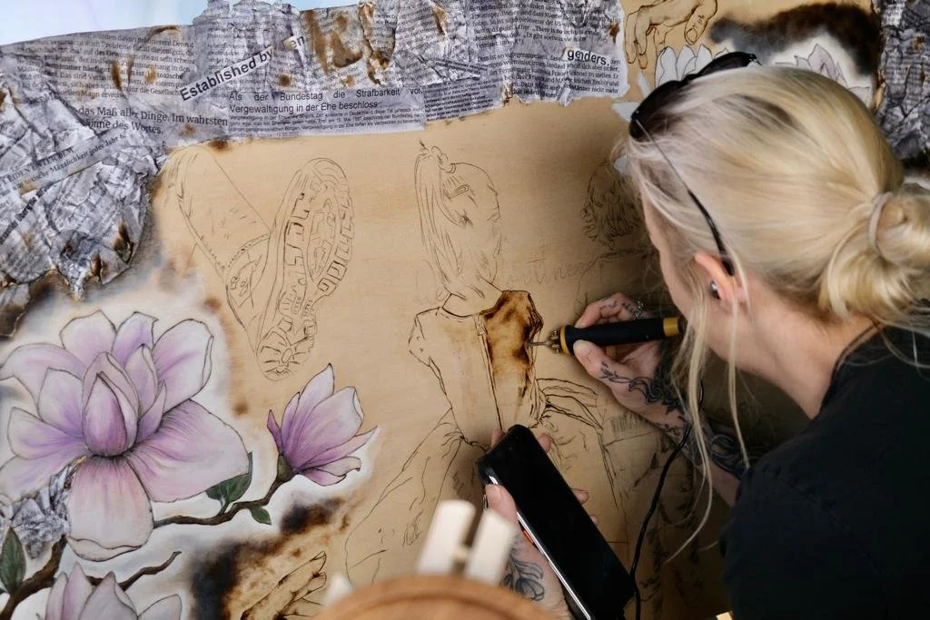 Frau mit blonden Haaren erstellt ein Kunstwerk mit einem Brennstab und einem Smartphone in der Hand. Auf der Leinwand befinden sich Zeichnungen von Blumen und einer Figur, umgeben von verbranntem Zeitungspapier. Das Kunstwerk hat Details von Schuhen und Blumen.