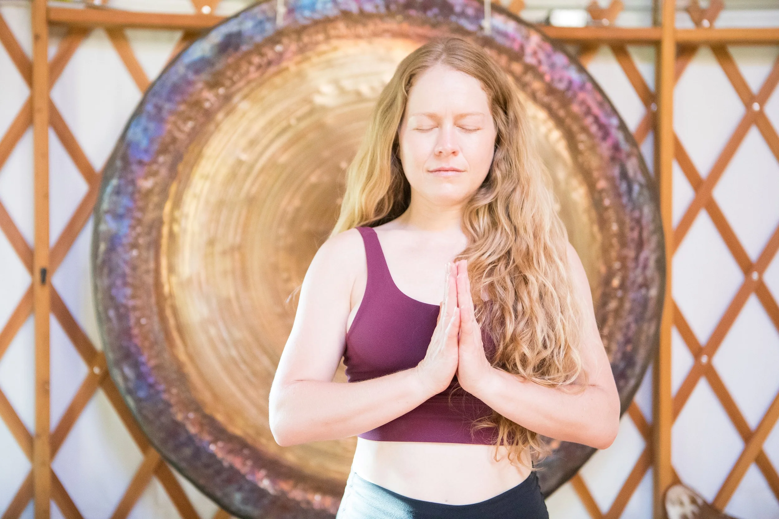 Woman with long red hair in a purple tank top practicing yoga or meditation in front of a large circular gong with a wooden lattice wall behind her.