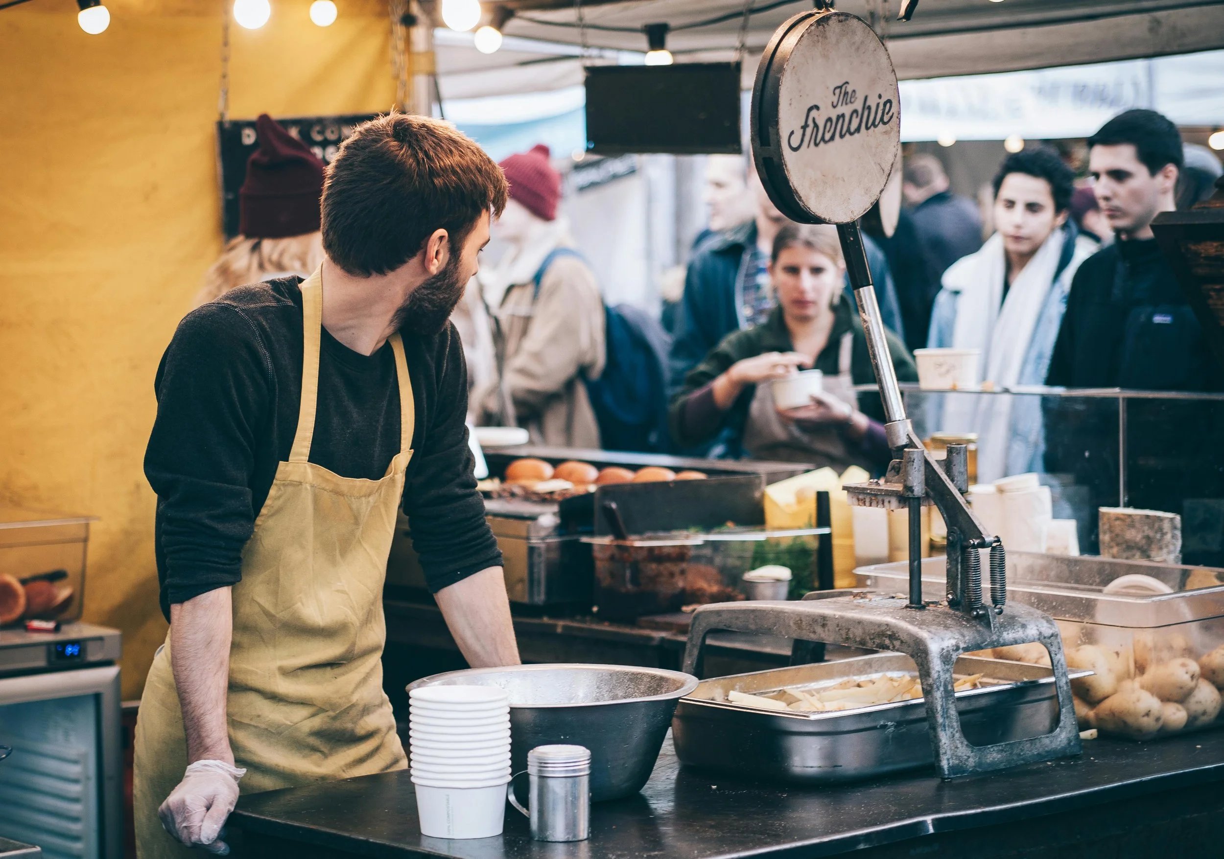 A man in a beige apron and black shirt working at a food stand with a vintage meat slicer, bowls, and containers, while a crowd of people waits in line at an outdoor market.