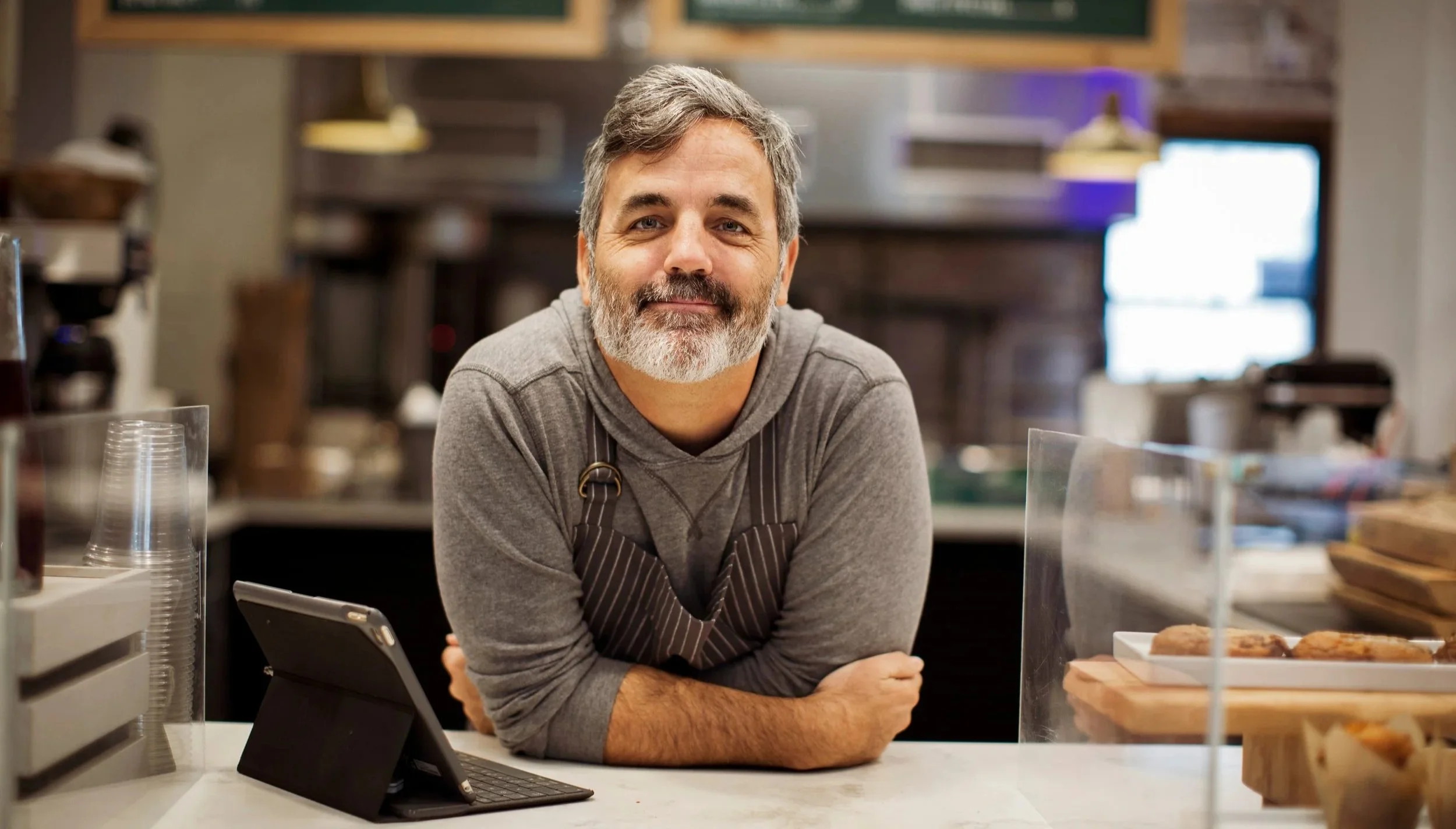 A friendly man with gray hair and a beard, wearing a gray hoodie and apron, leaning on a counter in a coffee shop or bakery with baked goods on display.