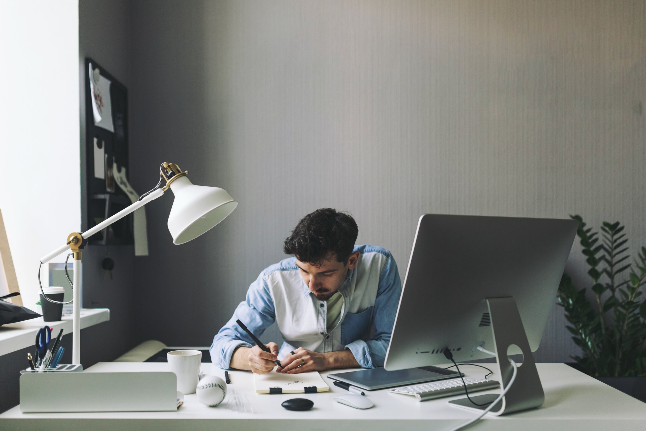 Man writing notes at a desk with computer, lamp, coffee cup, and office supplies in a modern office setting.