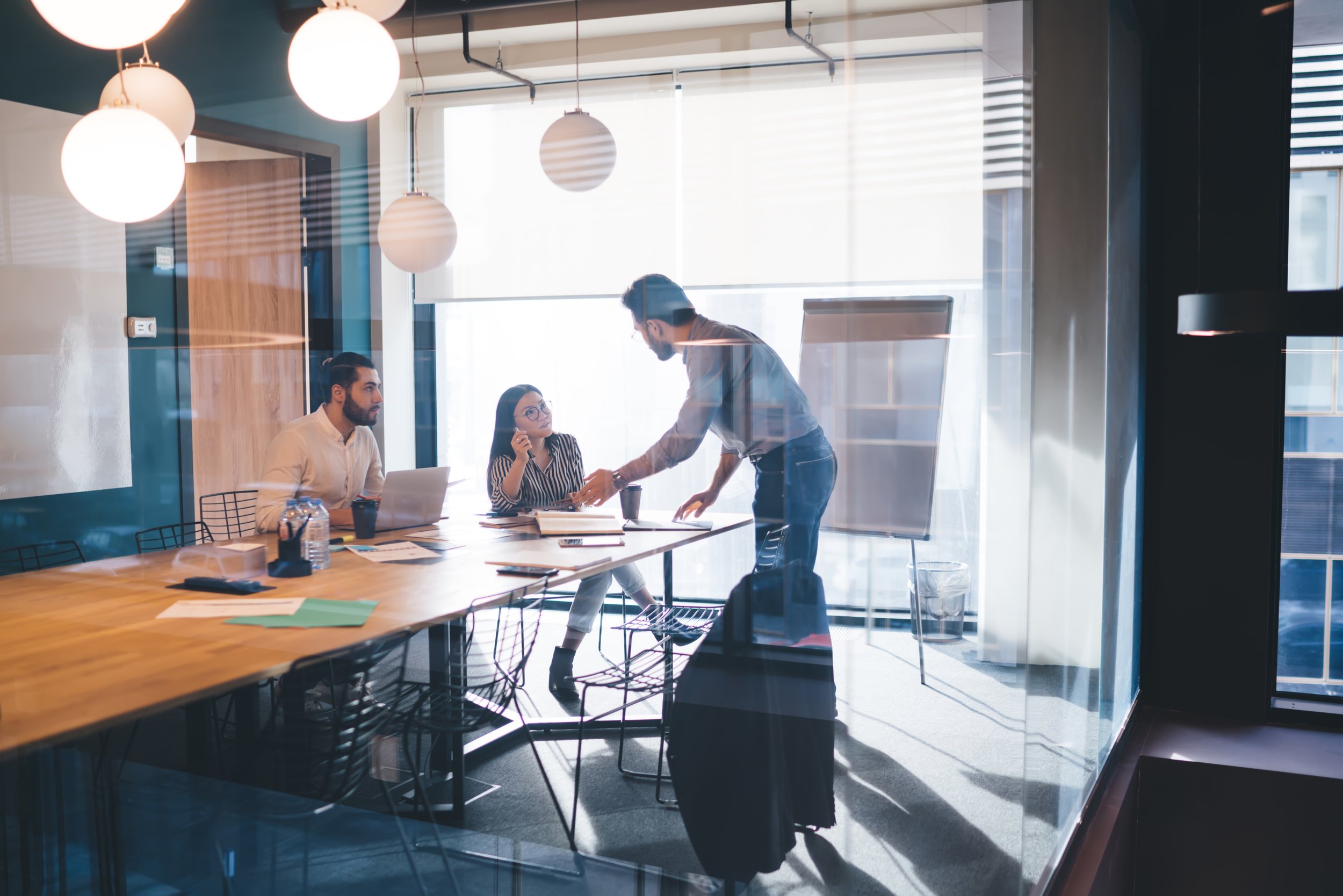 Three people in a modern office meeting room, two seated at a table and one standing, engaging in a discussion. The room has large windows with blinds, pendant lights overhead, and various office supplies on the table.