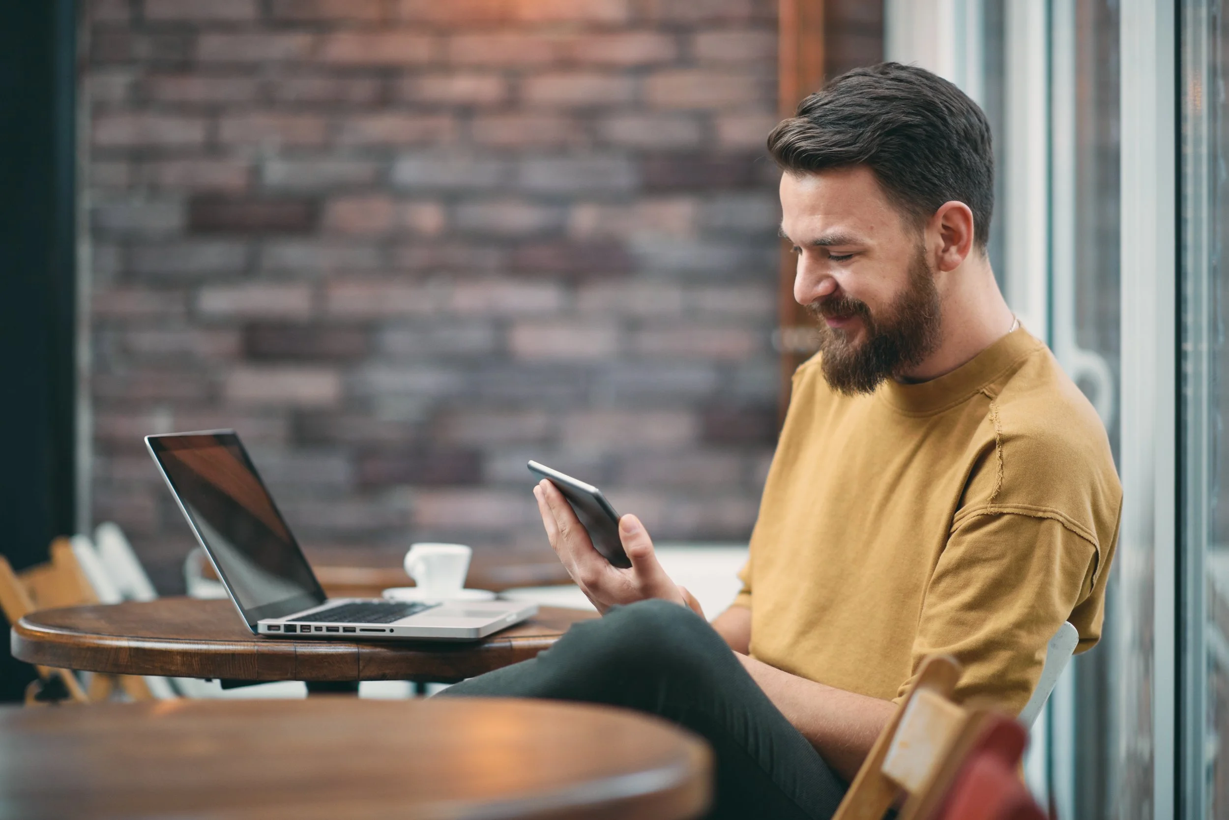 A man with a beard and short dark hair, wearing a yellow shirt, sitting at a wooden table in a cafe, looking at his smartphone with a smile. A laptop and a white cup are on the table.