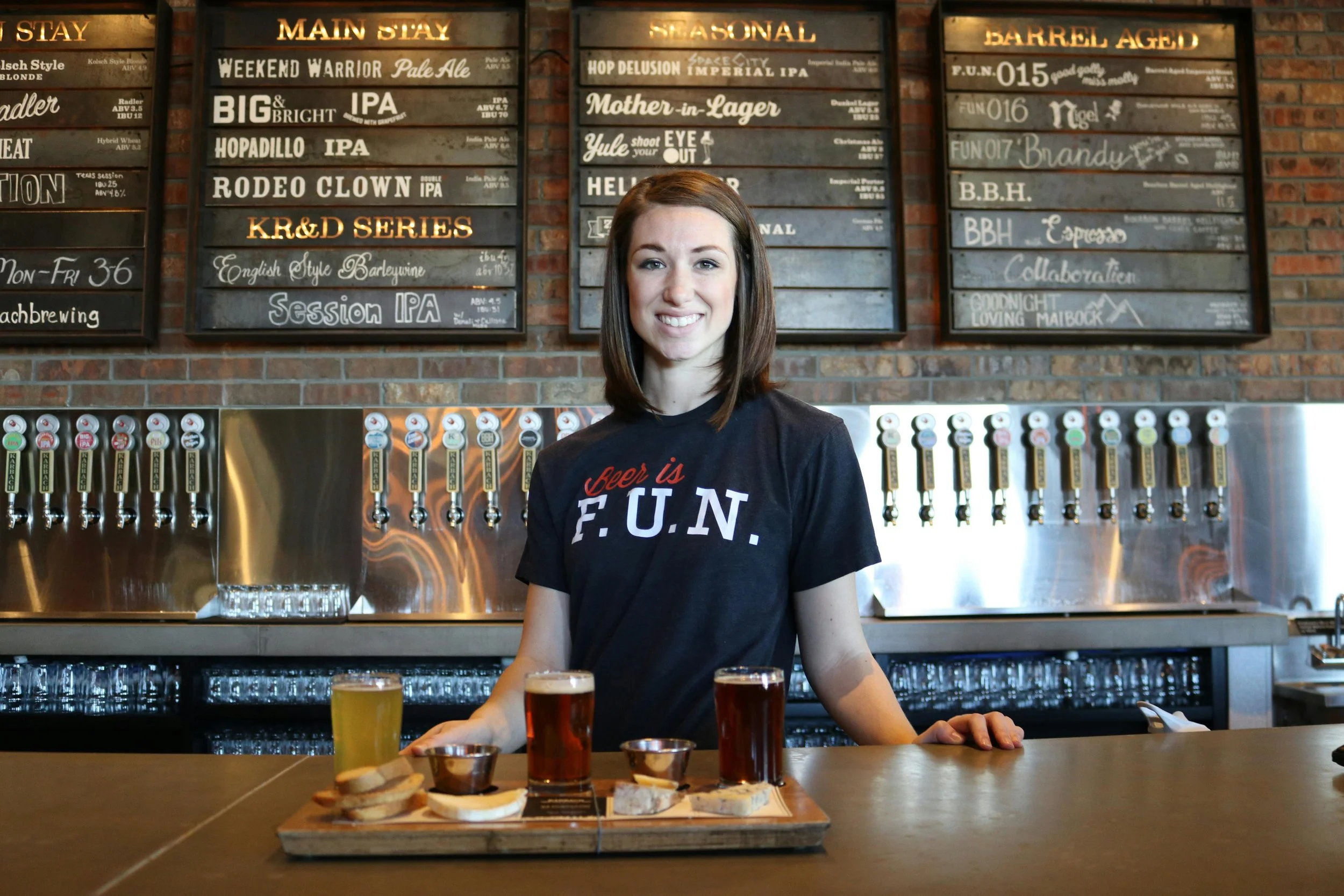 A woman standing behind a bar counter with three craft beers and a tasting platter, smiling at the camera with beer taps and menu boards in the background.