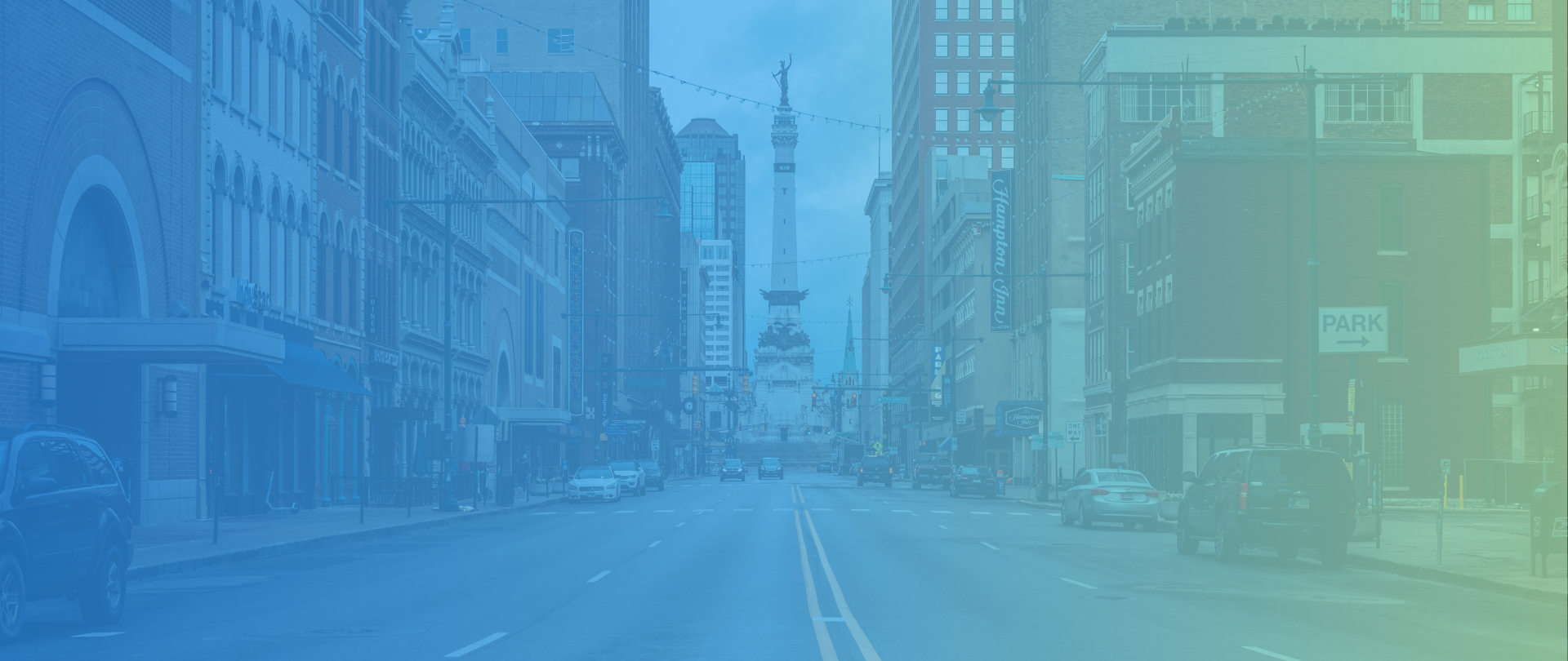 A city street scene with cars parked along the sides, a tall monument in the distance, and buildings lining the street, with a partly cloudy sky overhead.