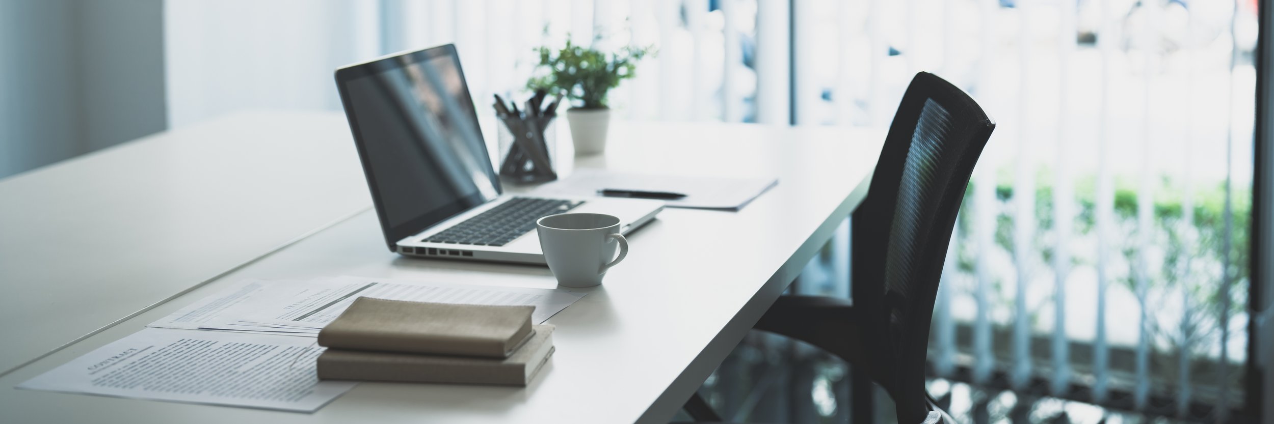 Office workspace with a white desk, open laptop, white coffee mug, stack of books, papers, pen, and a black office chair, near large windows with blinds and greenery outside.