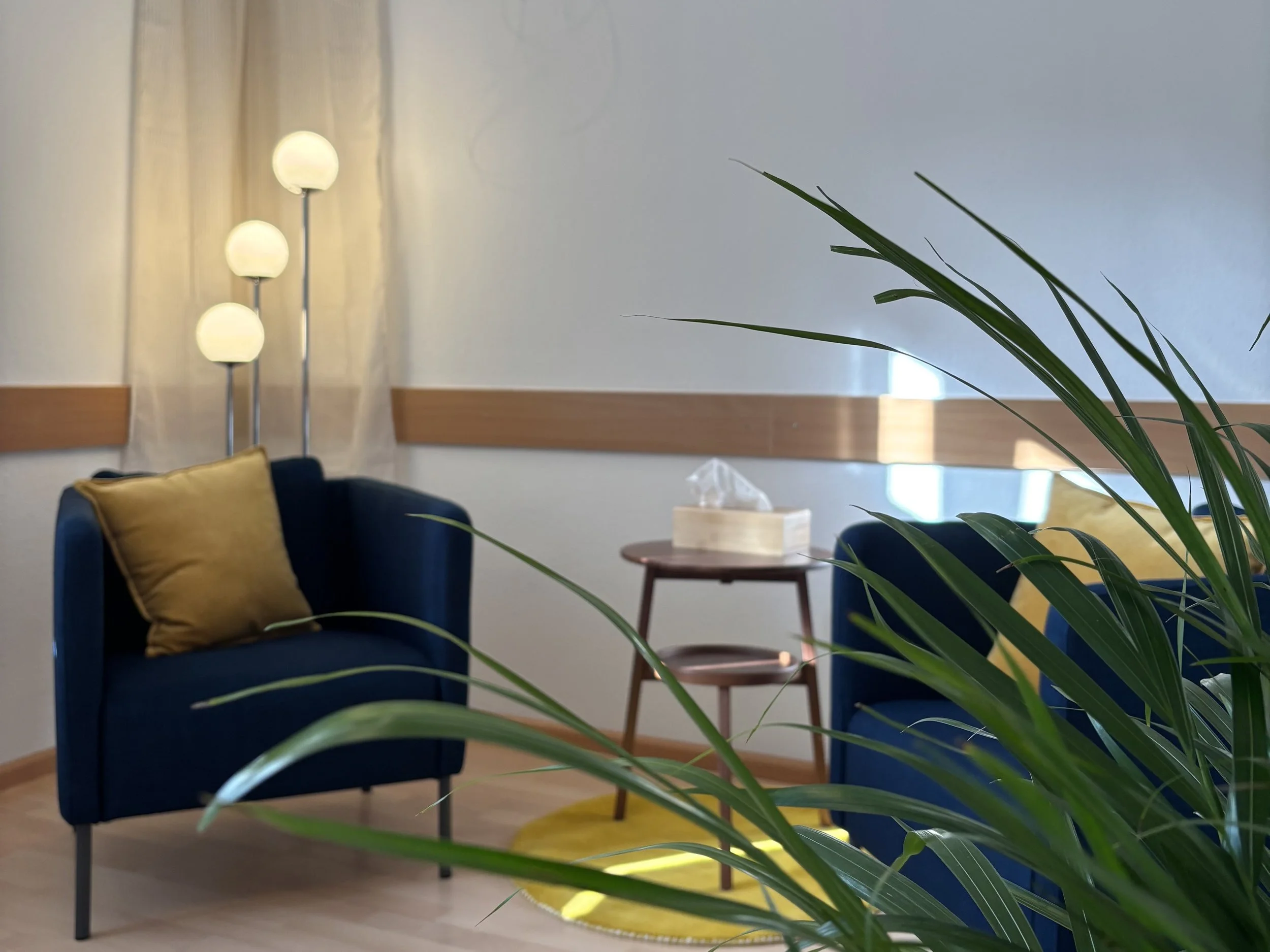 Gemütliches Wohnzimmer mit blauen Sesseln, gelben Kissen, holzfarbenem Boden, Tisch mit Papiertüchern, Pflanzen im Vordergrund, Stehlampe mit drei Kugeln, weiße Wände, Vorhänge