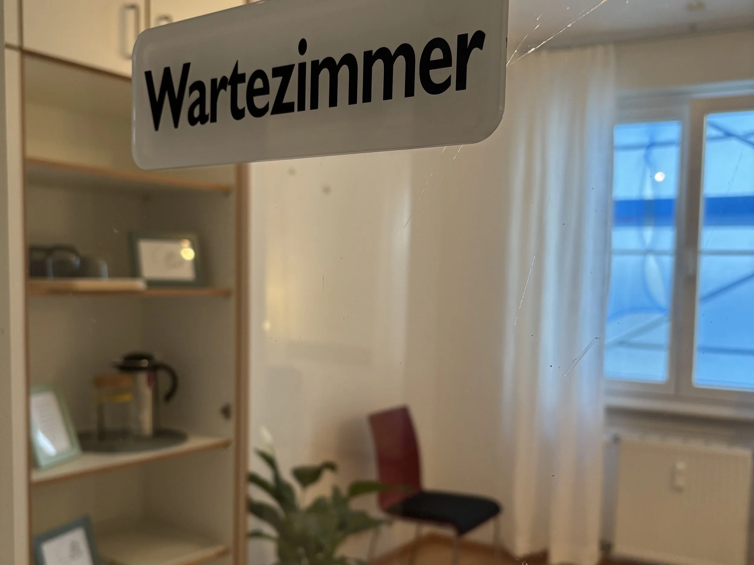 Innenraum eines Wartezimmers mit einem Stuhl, einem Fenster mit weißen Vorhängen und einem Regal. Es gibt auch einen kleinen Tisch mit einem Gerät darauf. Im Vordergrund ist eine Tür mit einem Schild "Wartezimmer".