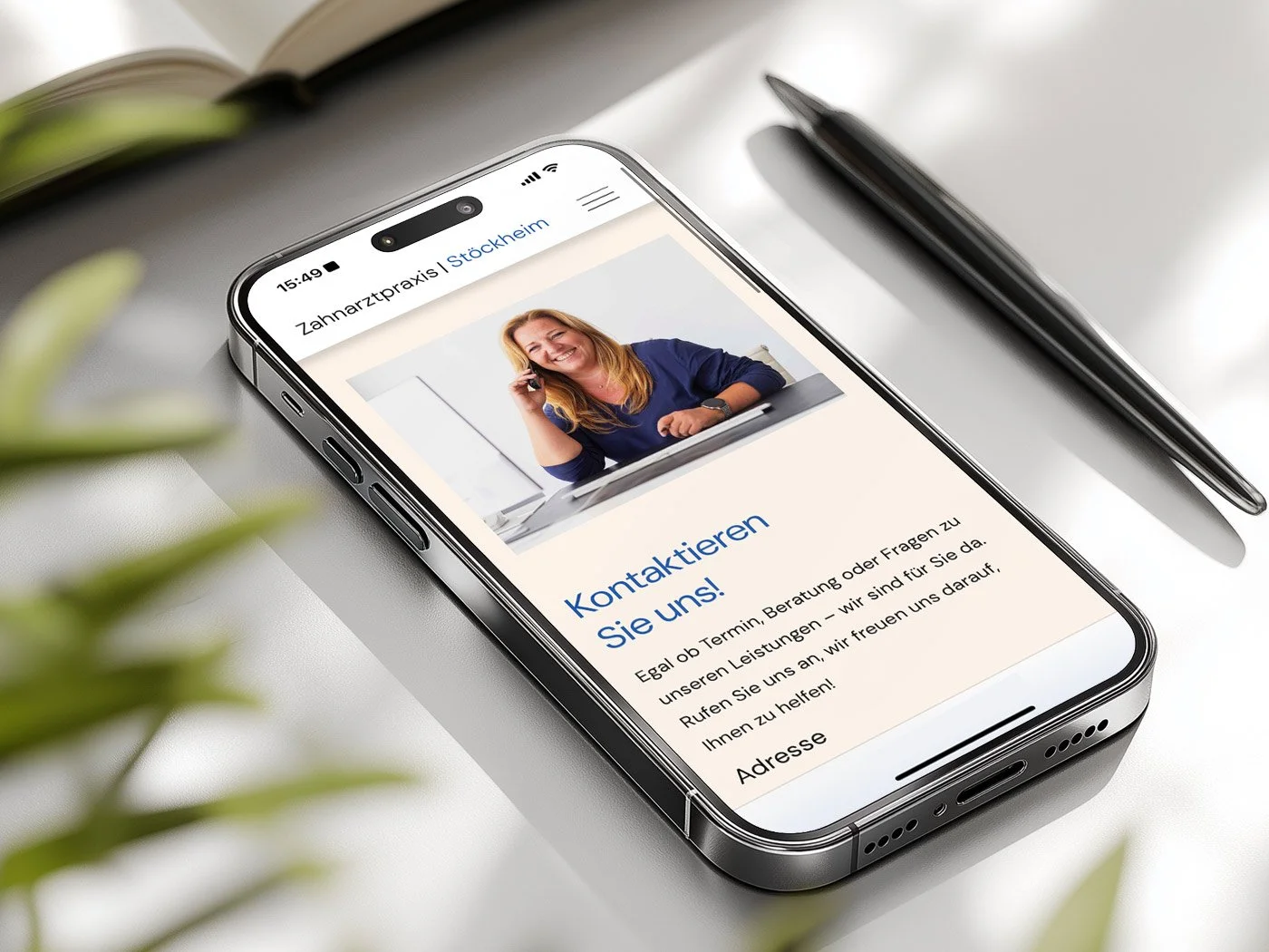 Smartphone auf einem Schreibtisch zeigt Webseite einer Zahnarztpraxis mit Bild einer lachenden Frau am Telefon, Text in deutscher Sprache. Umgeben von Notizbuch, Stift und Grünpflanzen.