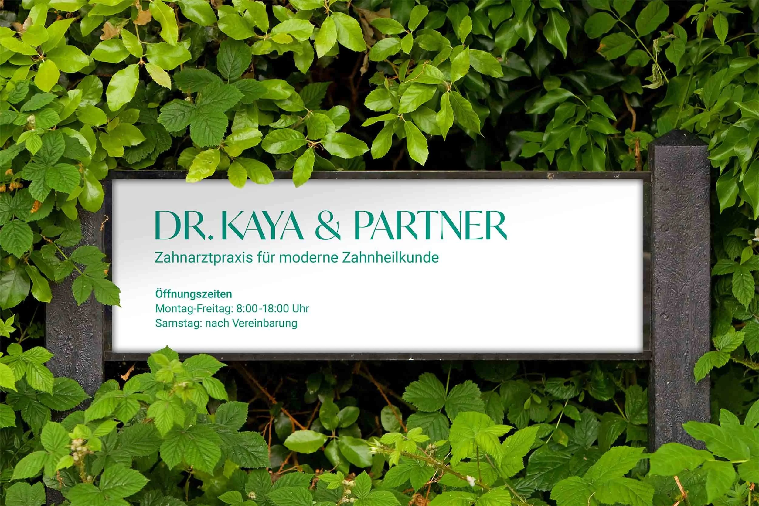 Praxisschild von Dr. Kaya & Partner in modernem Design mit Öffnungszeiten – eingebettet in grüne Pflanzenwand