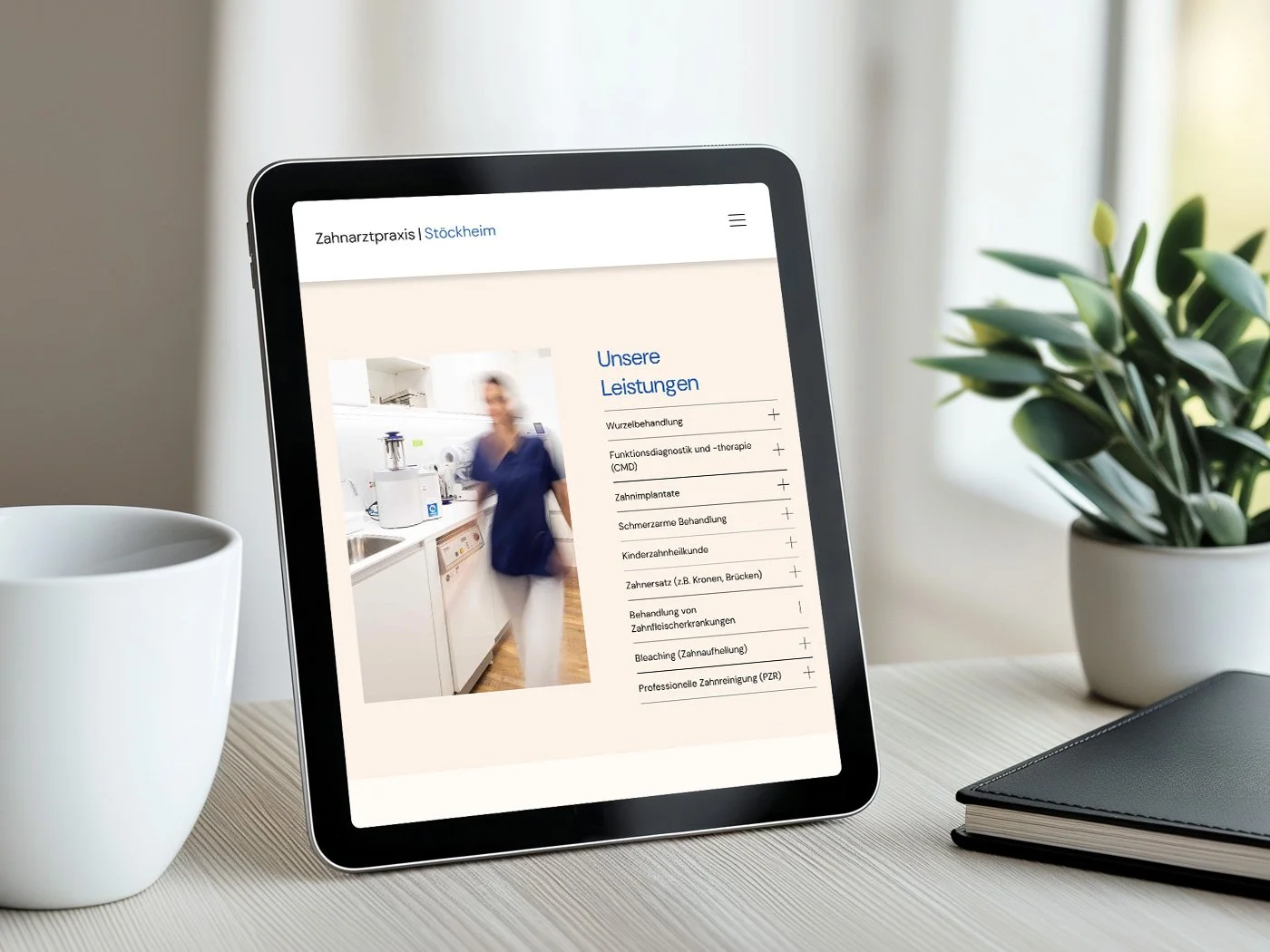 Tablet auf einem Schreibtisch zeigt Webseite einer Zahnarztpraxis mit Leistungsübersicht, neben einer weißen Teetasse, einem schwarzen Notizbuch und einer Topfpflanze.