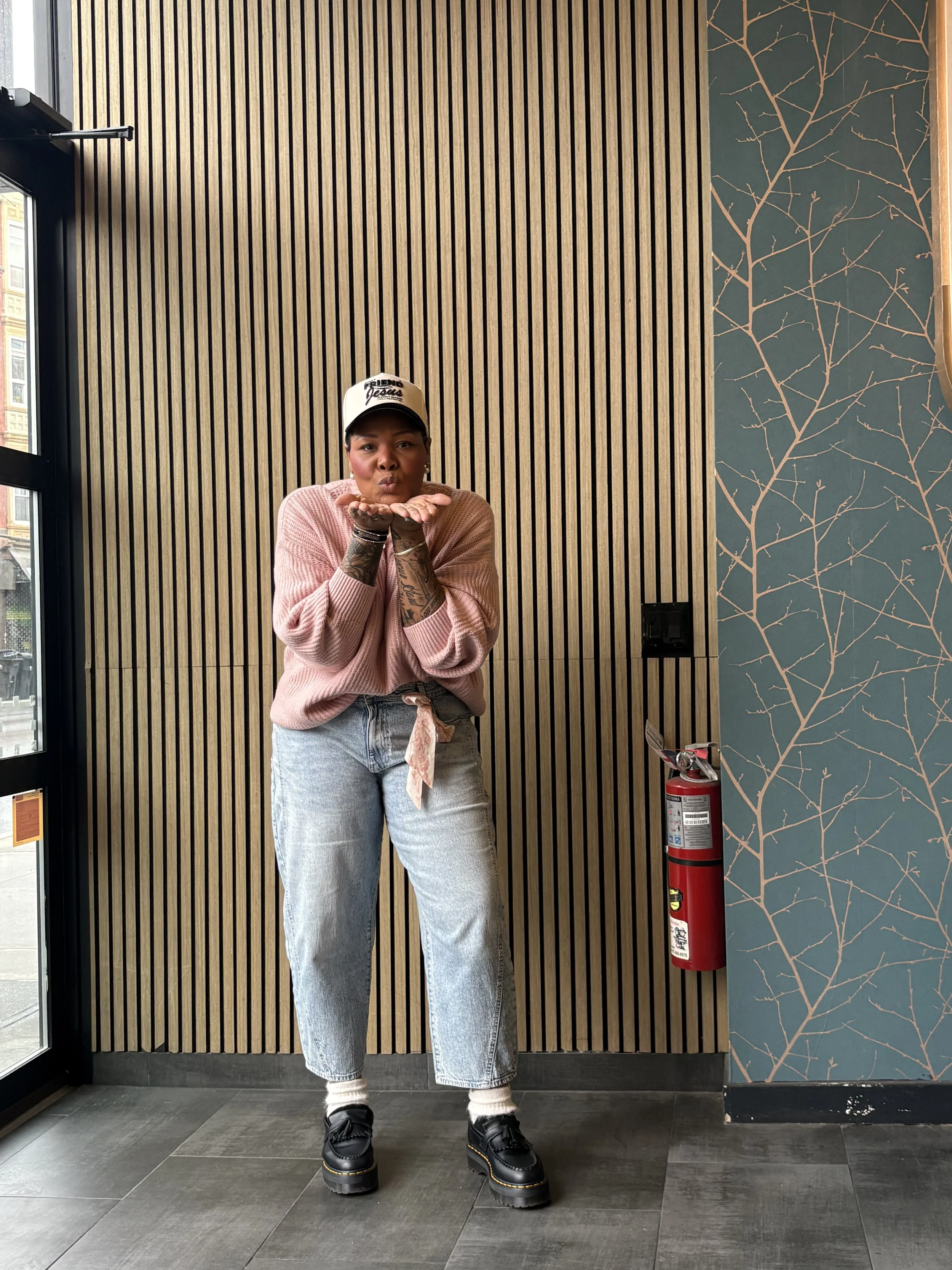 A woman standing indoors near a wall with vertical wooden slats and a blue wall with a branch pattern, blowing a kiss and wearing casual clothing, a cap, and chunky black shoes.