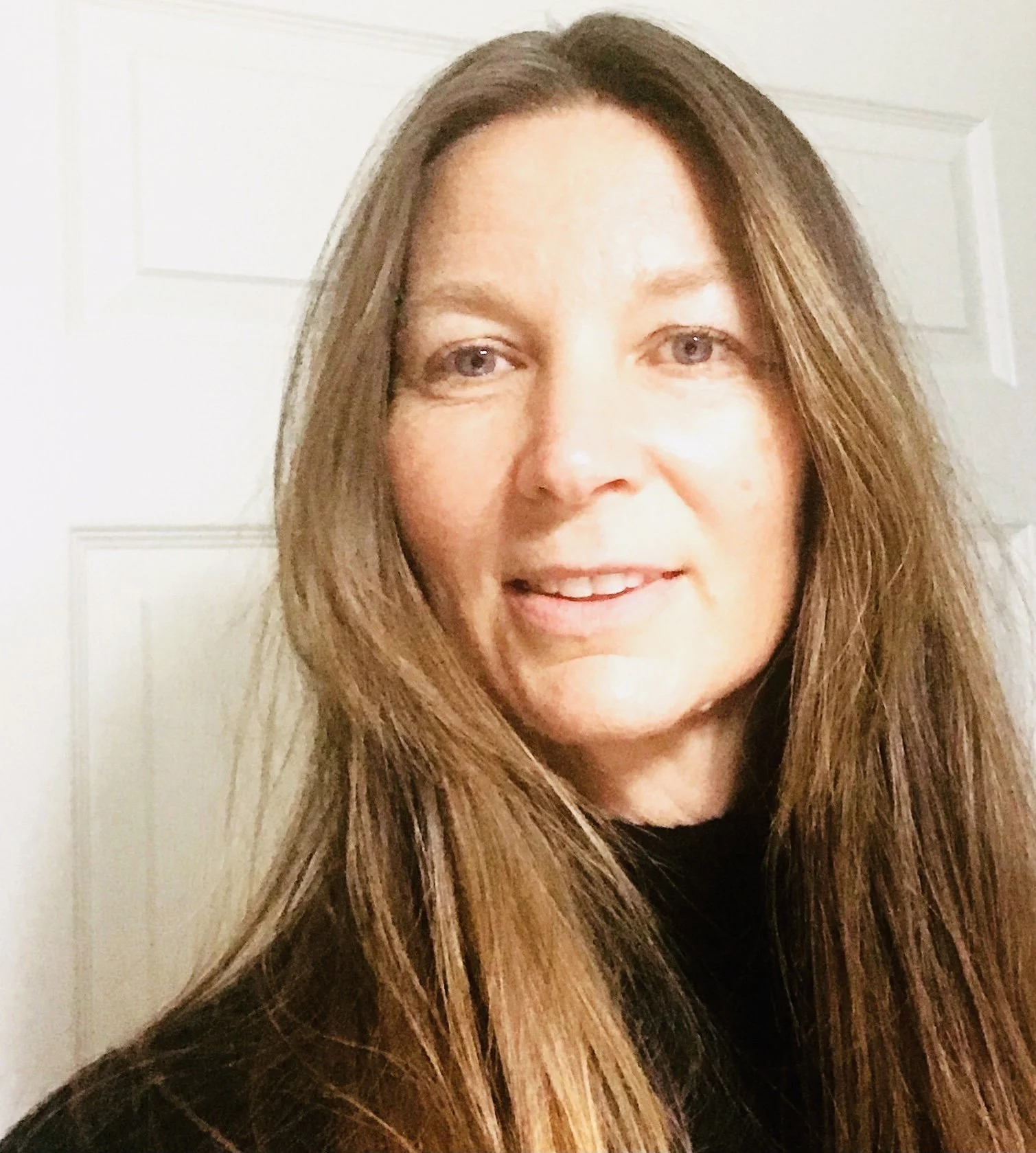 Close-up of a woman with long brown hair smiling, standing in front of a paneled white door.