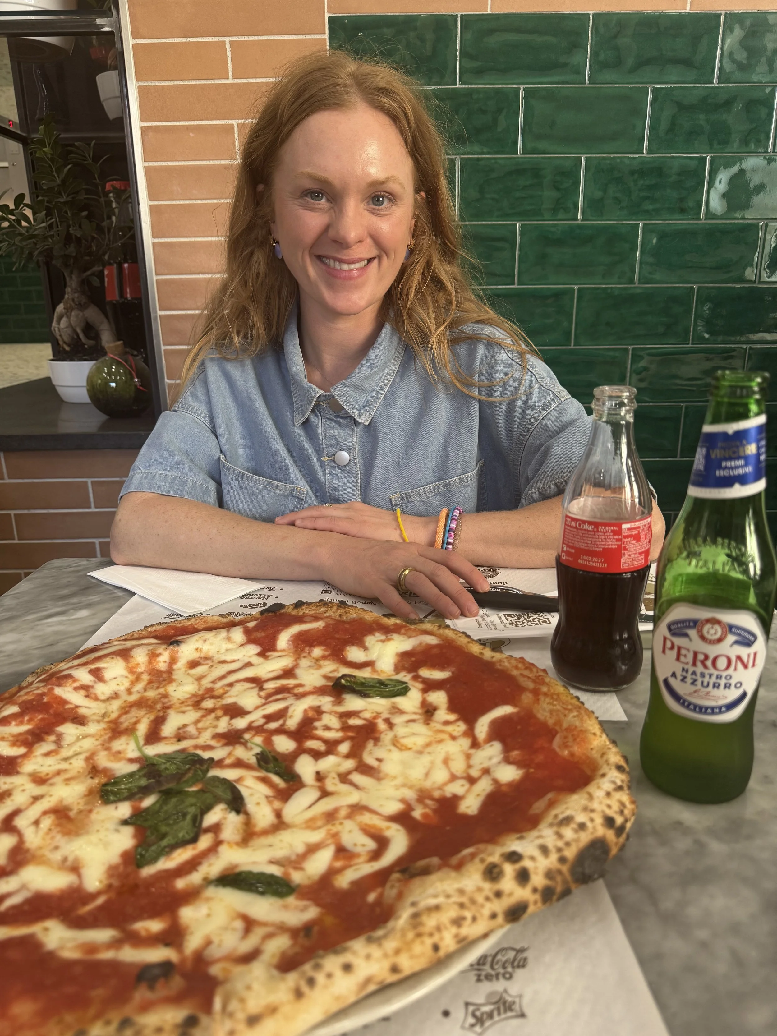 A women with long strawberry blonde hair, in enjoying pizza and a refreshing beverage. Green tile and brick in the background. 